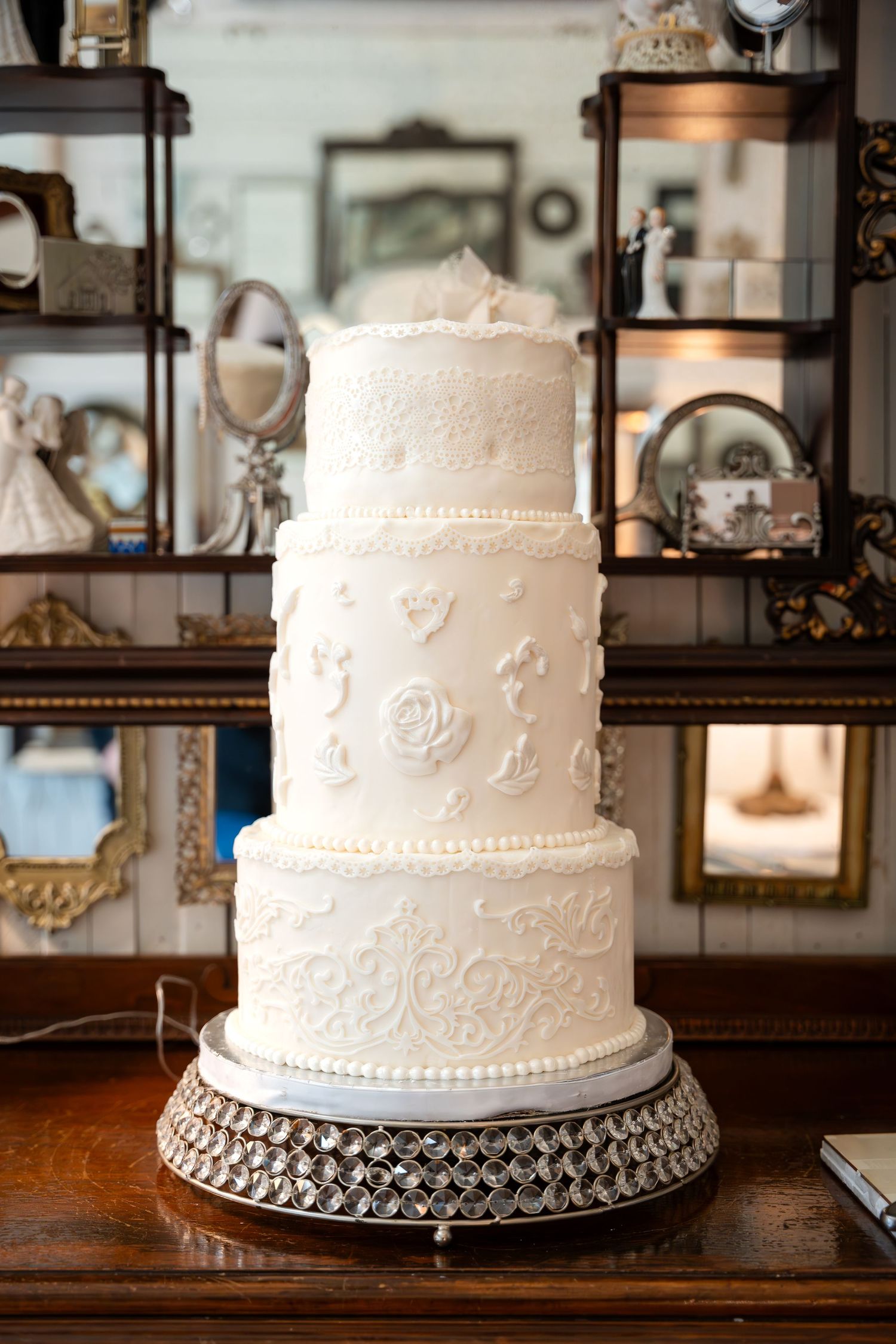Elegant three-tiered white wedding cake with delicate floral details on an ornate silver stand displayed against vintage decor.