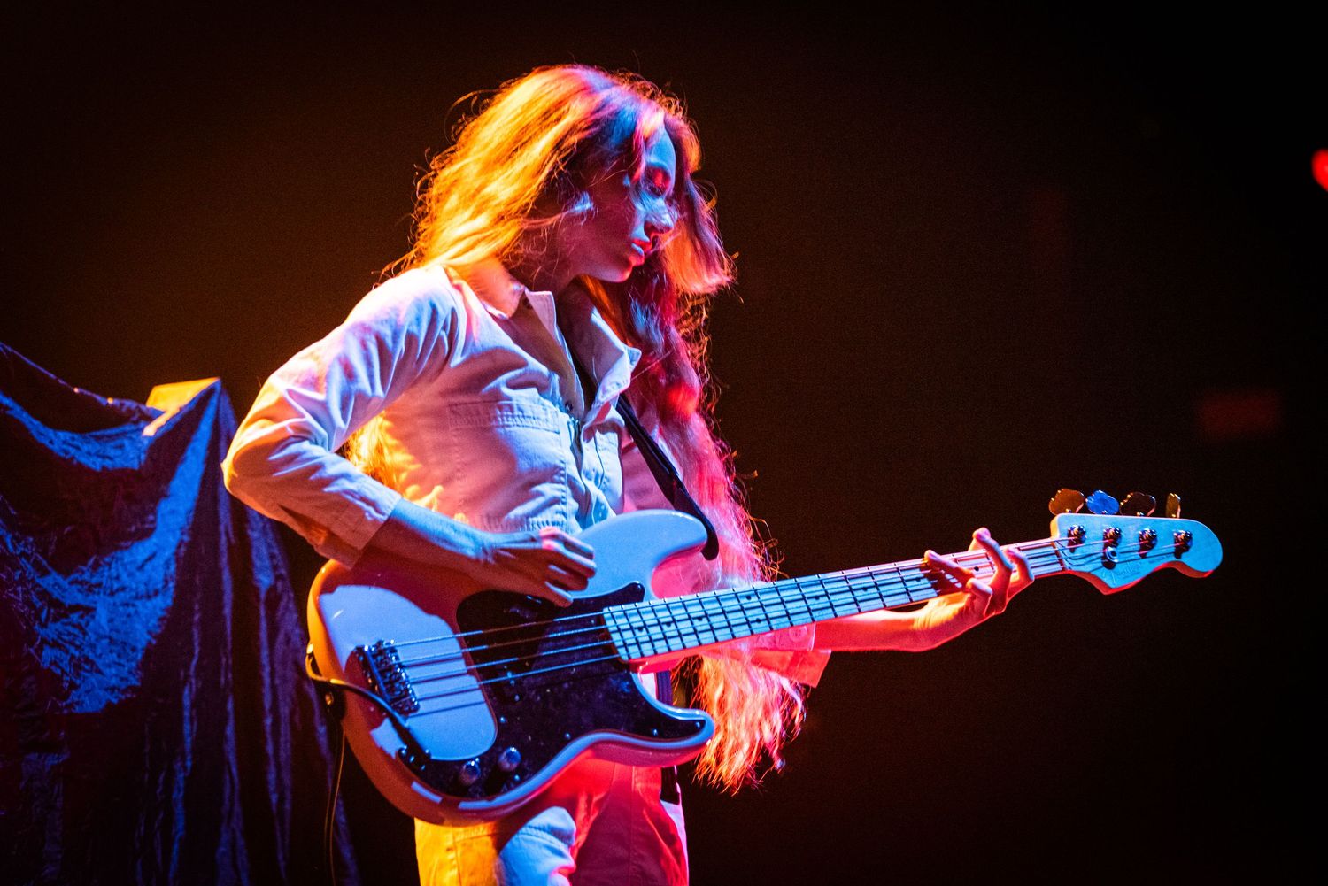 Bass guitarist performing under red and blue stage lighting with long hair flowing.