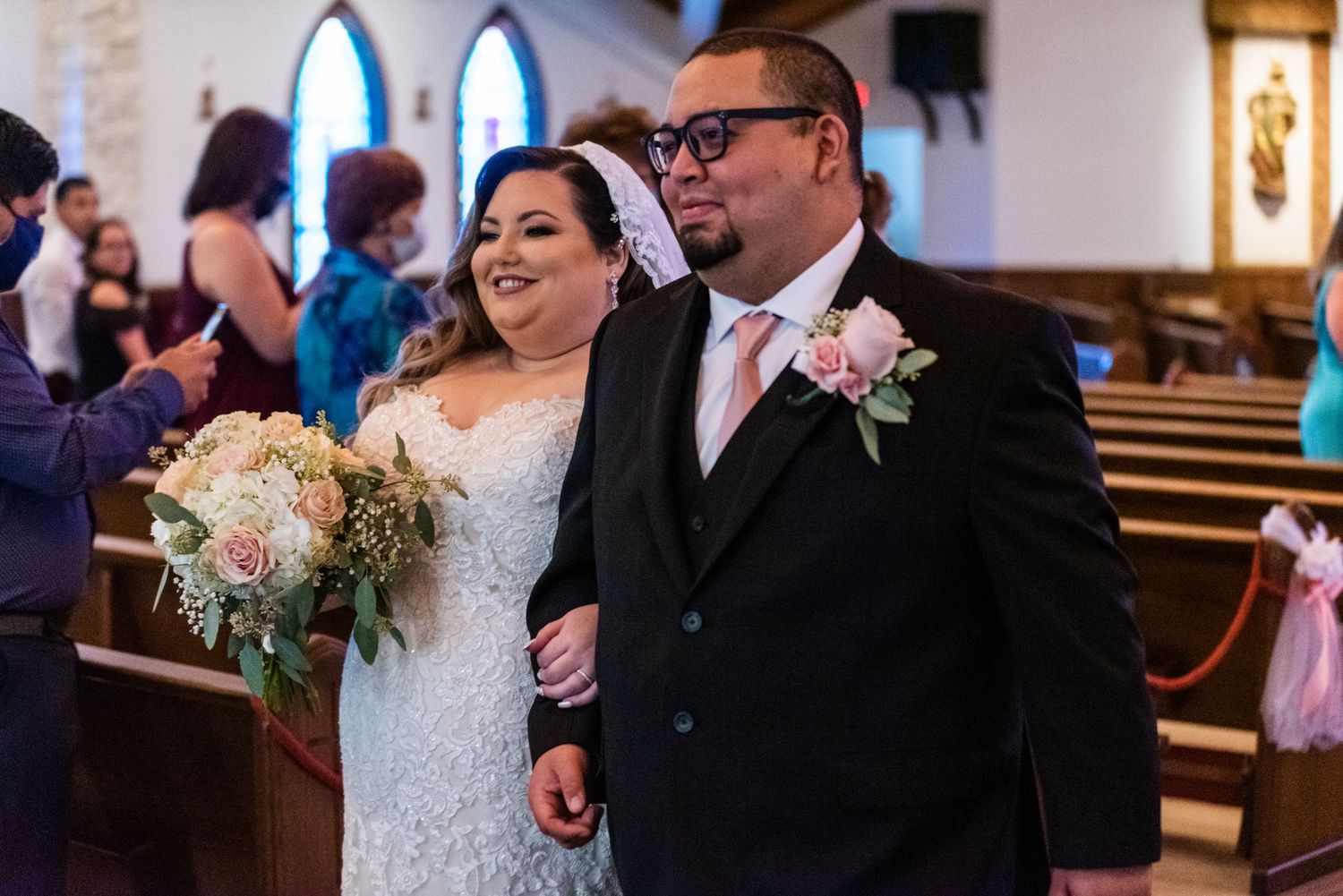 Happy newlyweds celebrate while walking down church aisle with bouquet and formal attire.