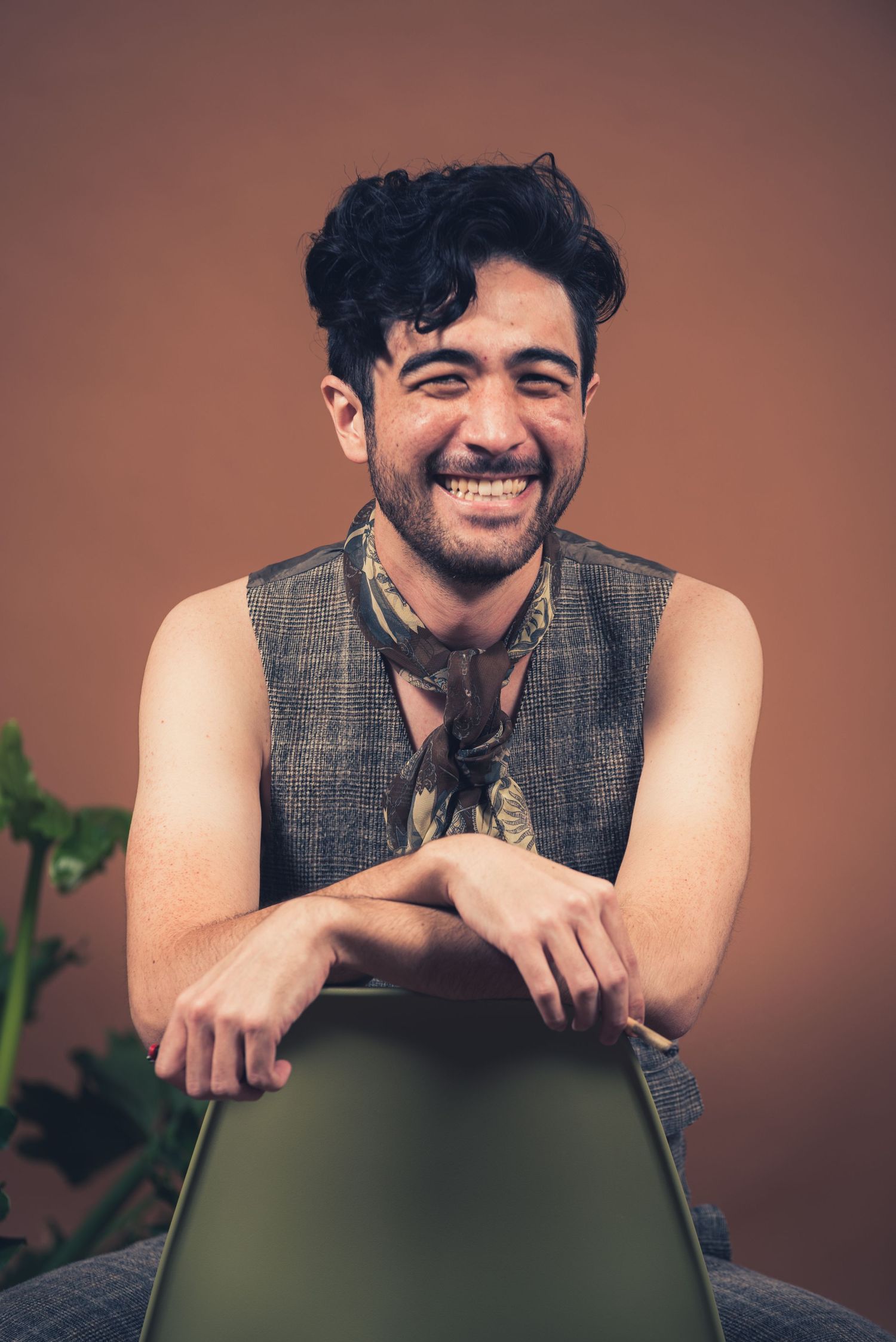 Person in grey vest leans on chair with joyful expression against brown studio backdrop.
