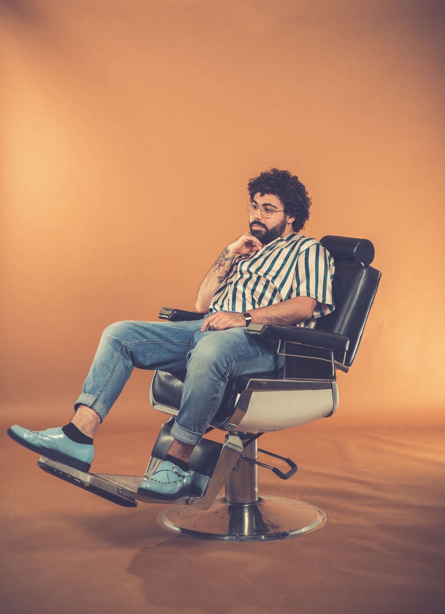 Person in striped shirt sitting casually in vintage barber chair against orange background.