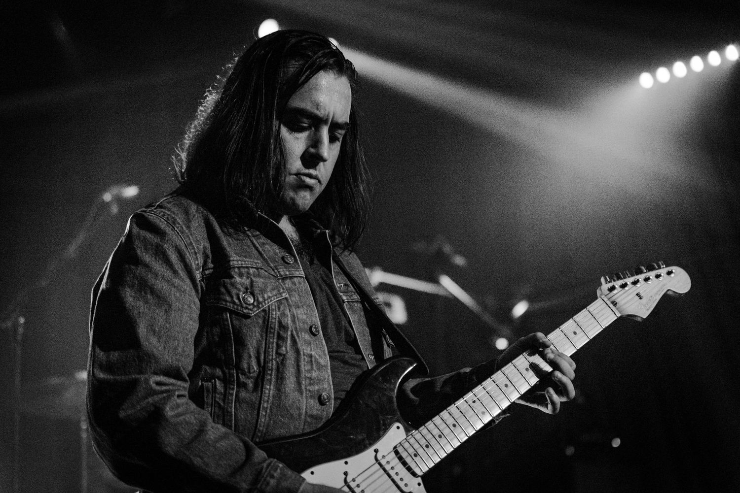 A guitarist in a denim jacket plays on stage under spotlights.