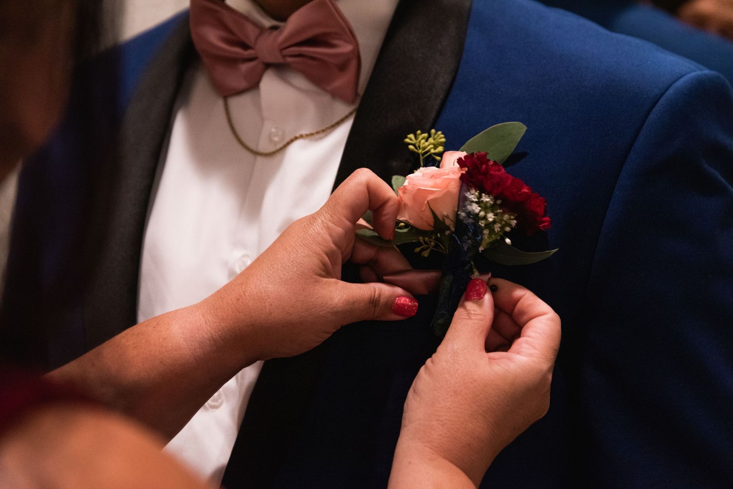 Hands pinning a boutonniere with red and pink flowers onto a navy blue suit jacket.