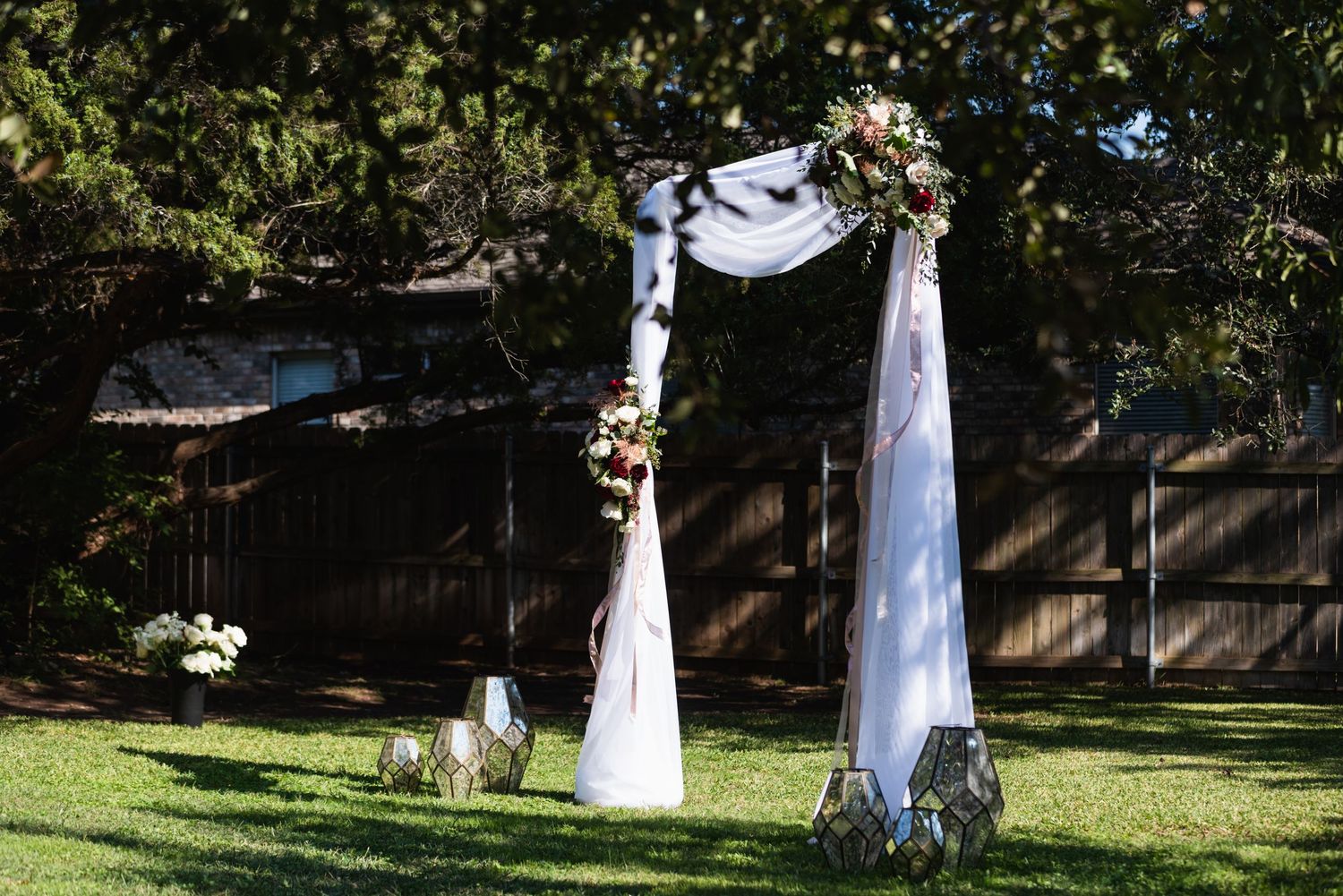 White fabric wedding arch with floral decorations stands in a sunlit backyard garden setting.