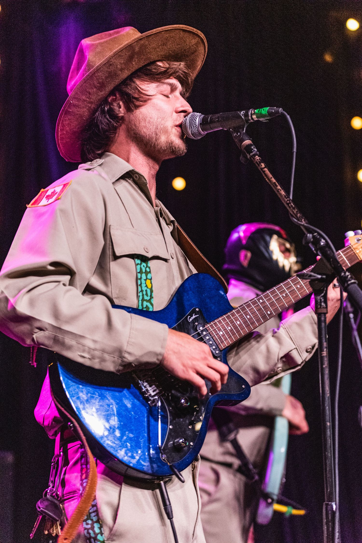 Performer in western attire plays blue guitar and sings into microphone on stage with purple lighting.