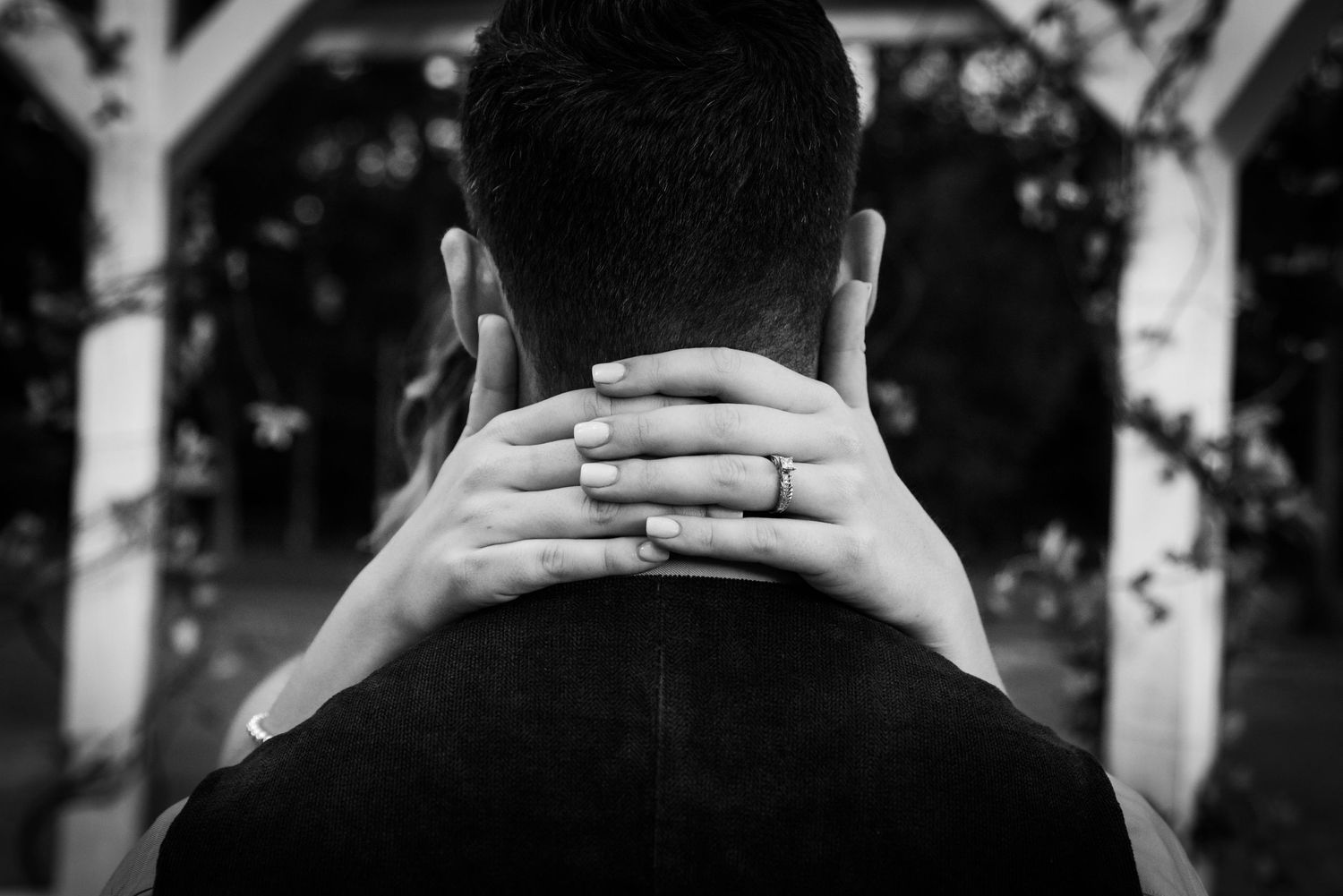 Delicate hands wearing a wedding ring wrap around a person's neck in black and white.