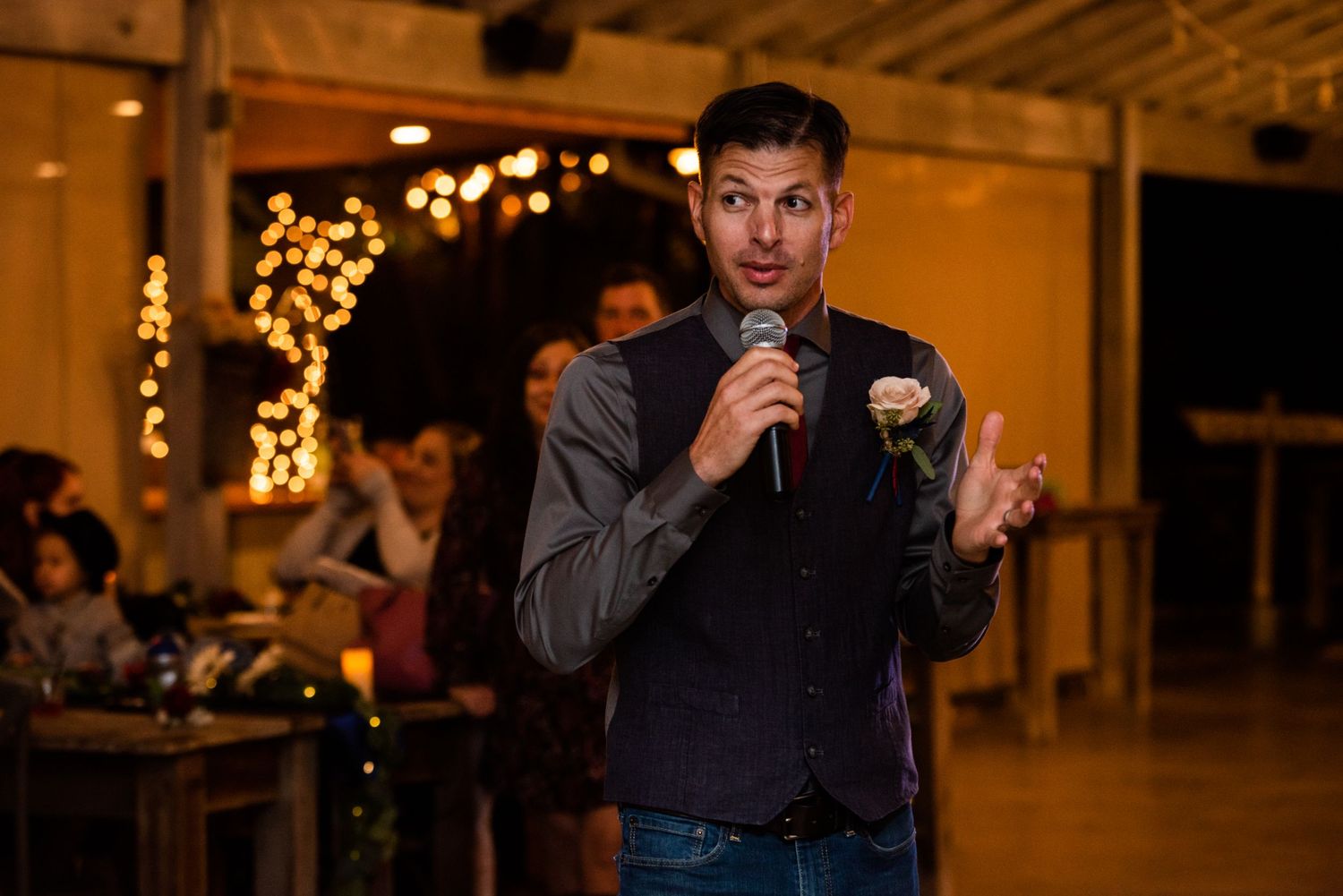 Speaker addresses wedding guests in industrial venue decorated with string lights.