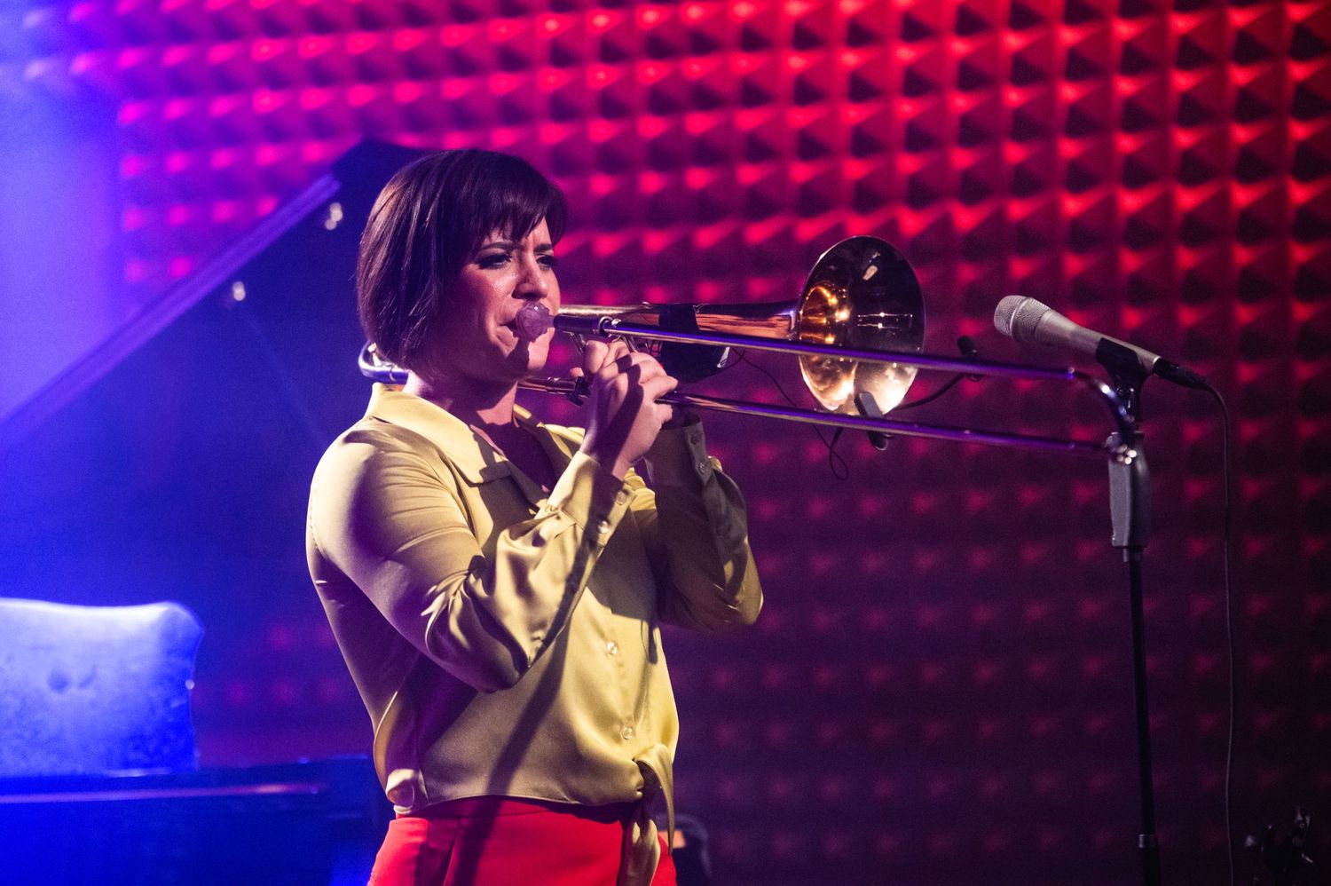 A performer plays trombone while wearing a yellow top against a red-lit textured background.