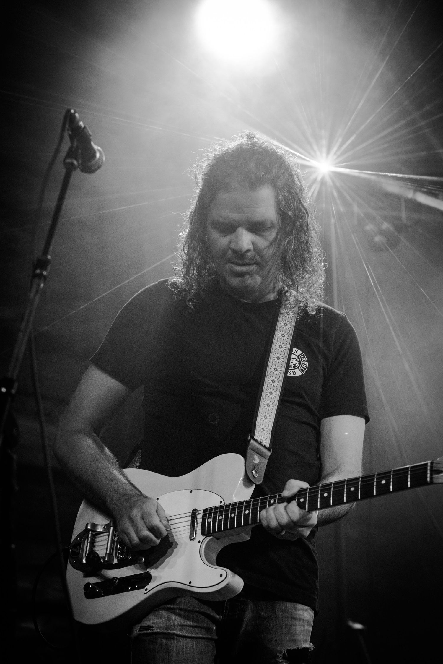 A guitarist plays a white electric guitar under dramatic stage spotlights in black and white.