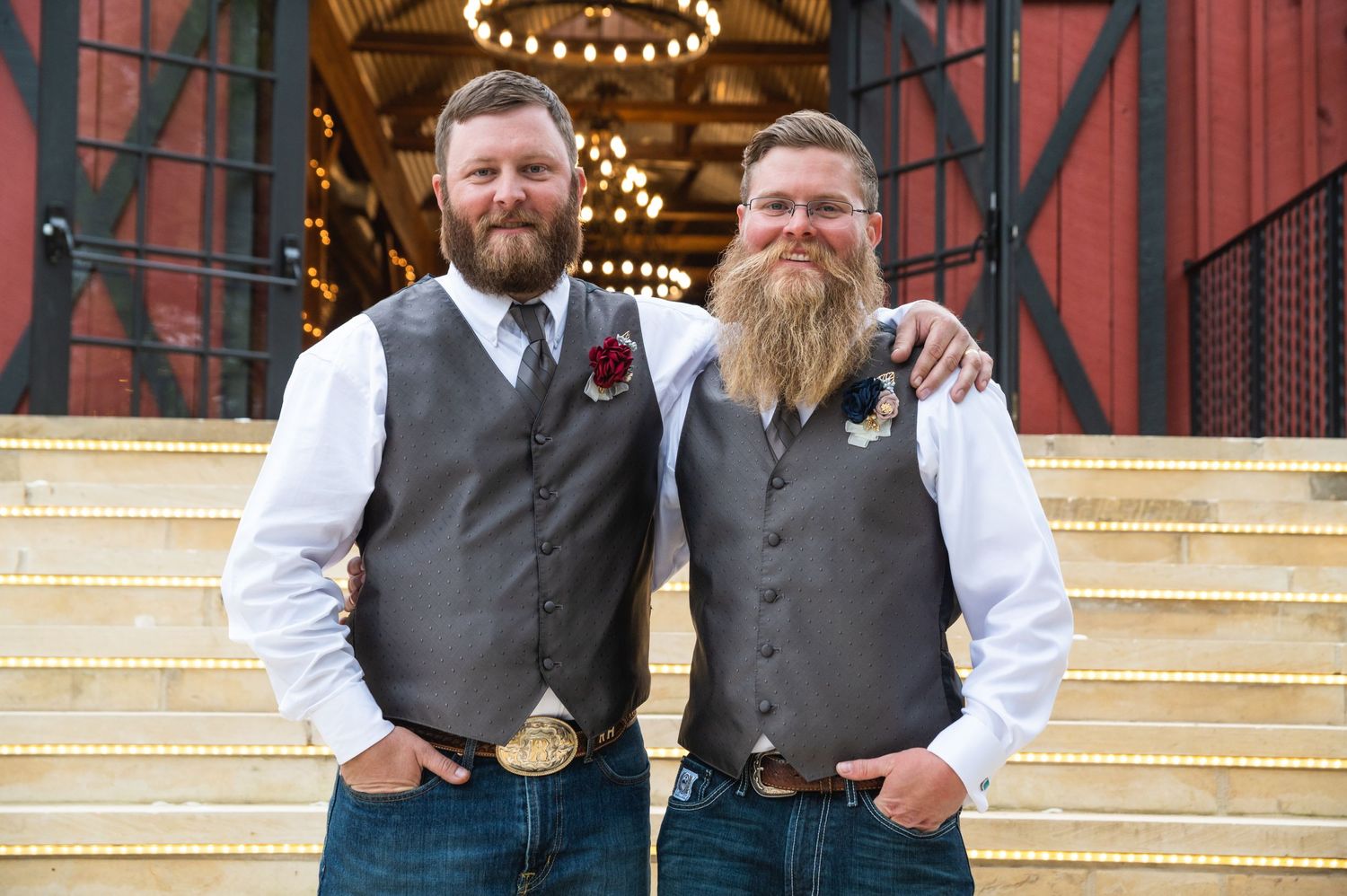Two groomsmen in matching gray vests pose on barn steps featuring impressive beards.