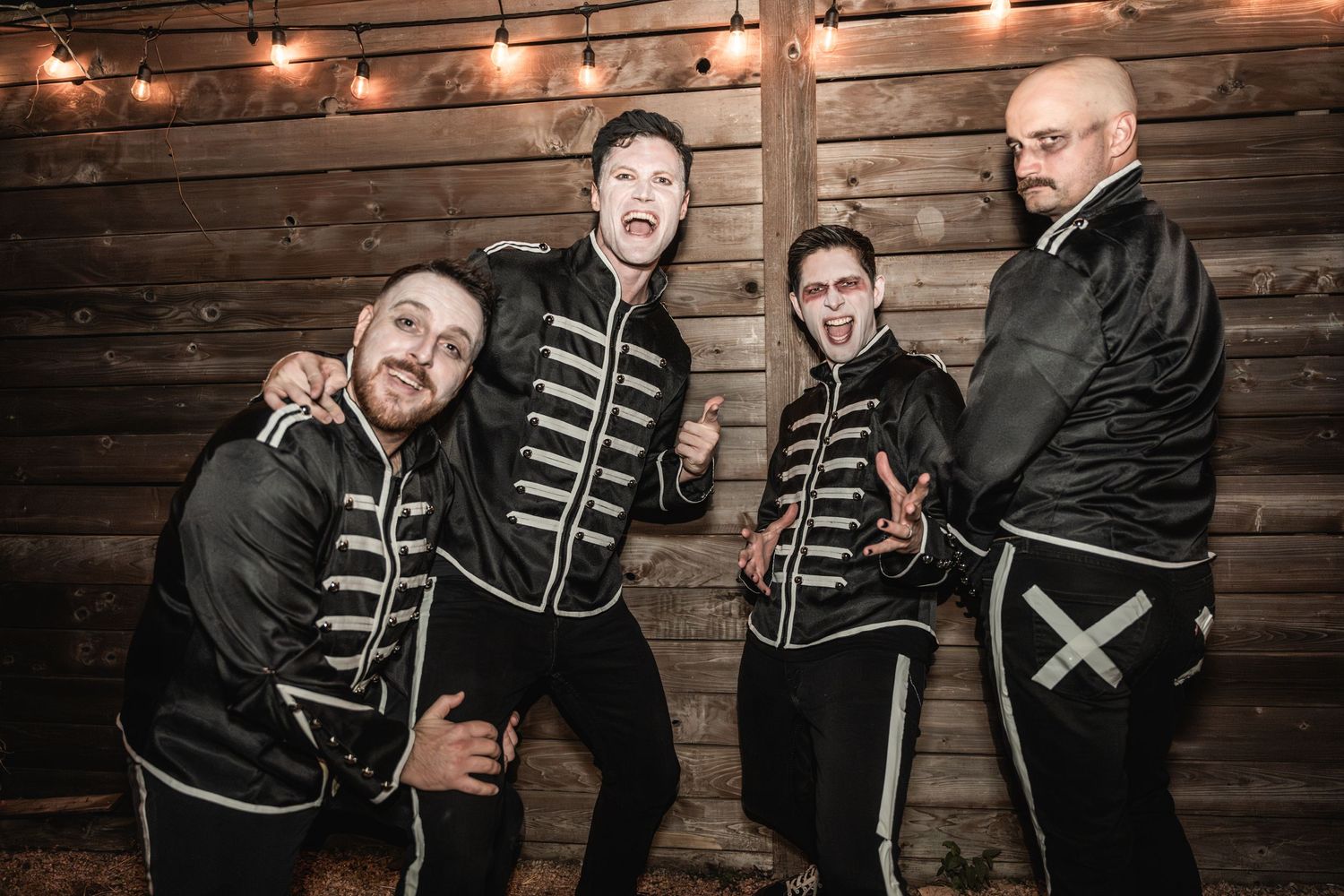 Band members strike dynamic poses in military-style jackets against rustic wooden backdrop.