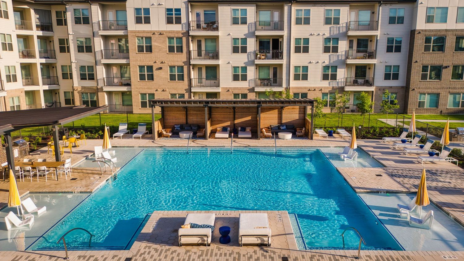 Resort-style apartment pool area with pergola and yellow umbrellas on a sunny day.