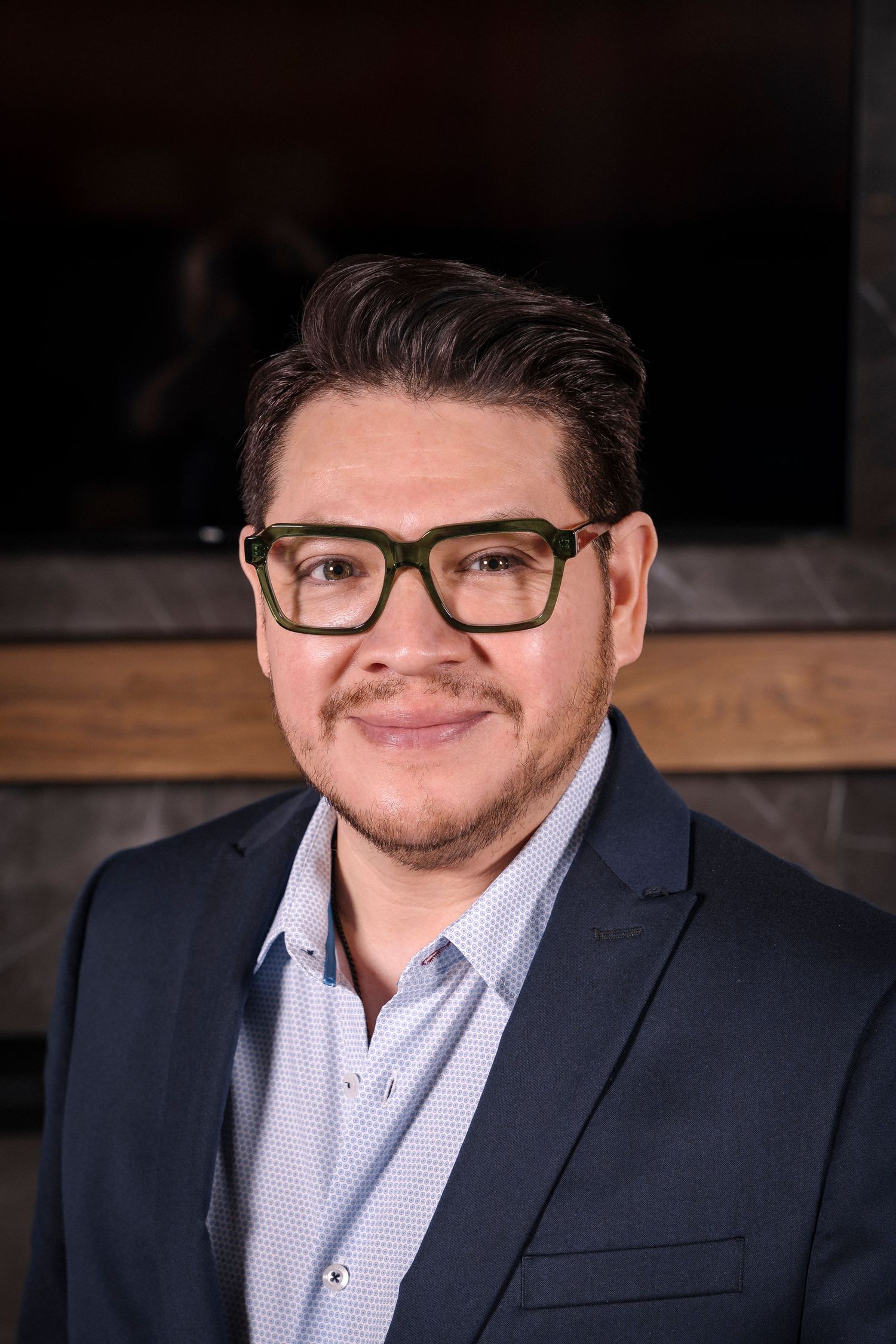 Professional headshot of person in navy suit jacket and glasses smiling at camera against dark background.