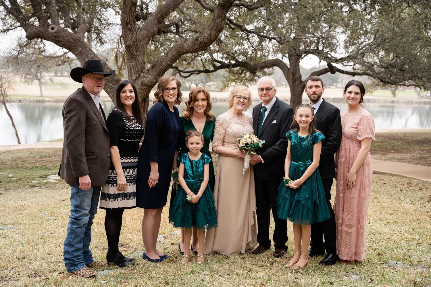 Wedding party poses outdoors under oak trees wearing emerald green and mauve dresses for rustic country celebration.