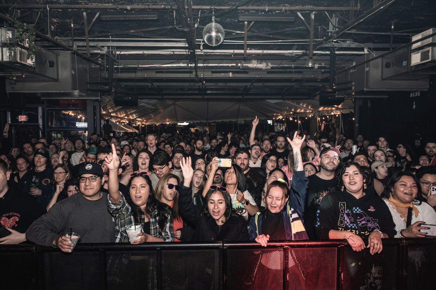 An enthusiastic concert crowd packs a dark venue with raised hands and excited expressions behind a security barrier.