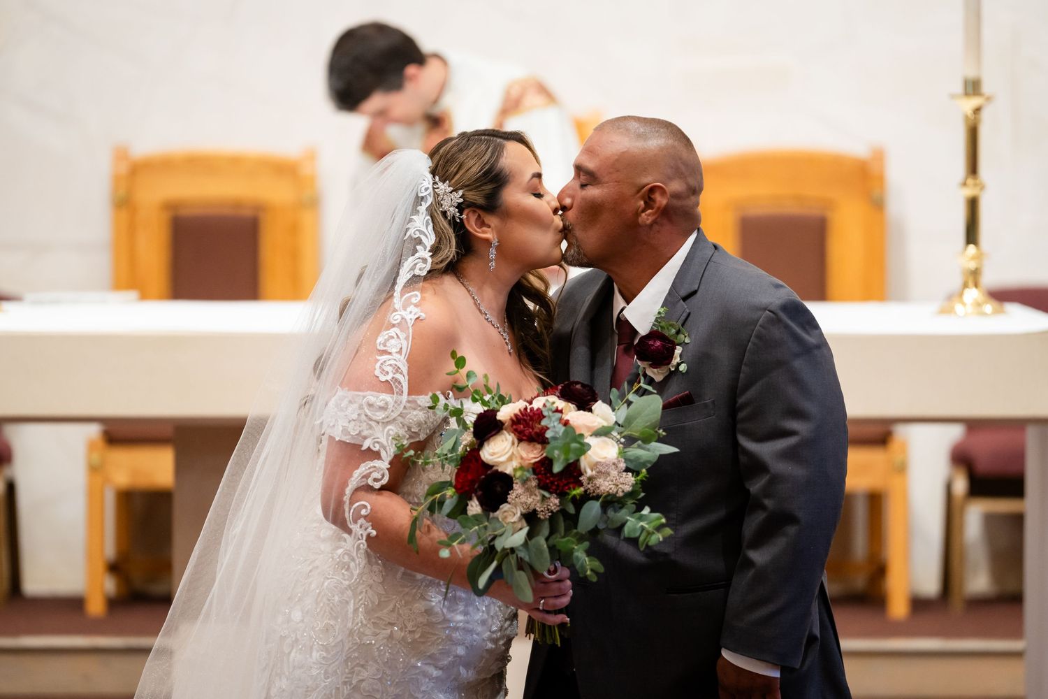 Newlyweds share romantic moment with burgundy flower bouquet during church ceremony.