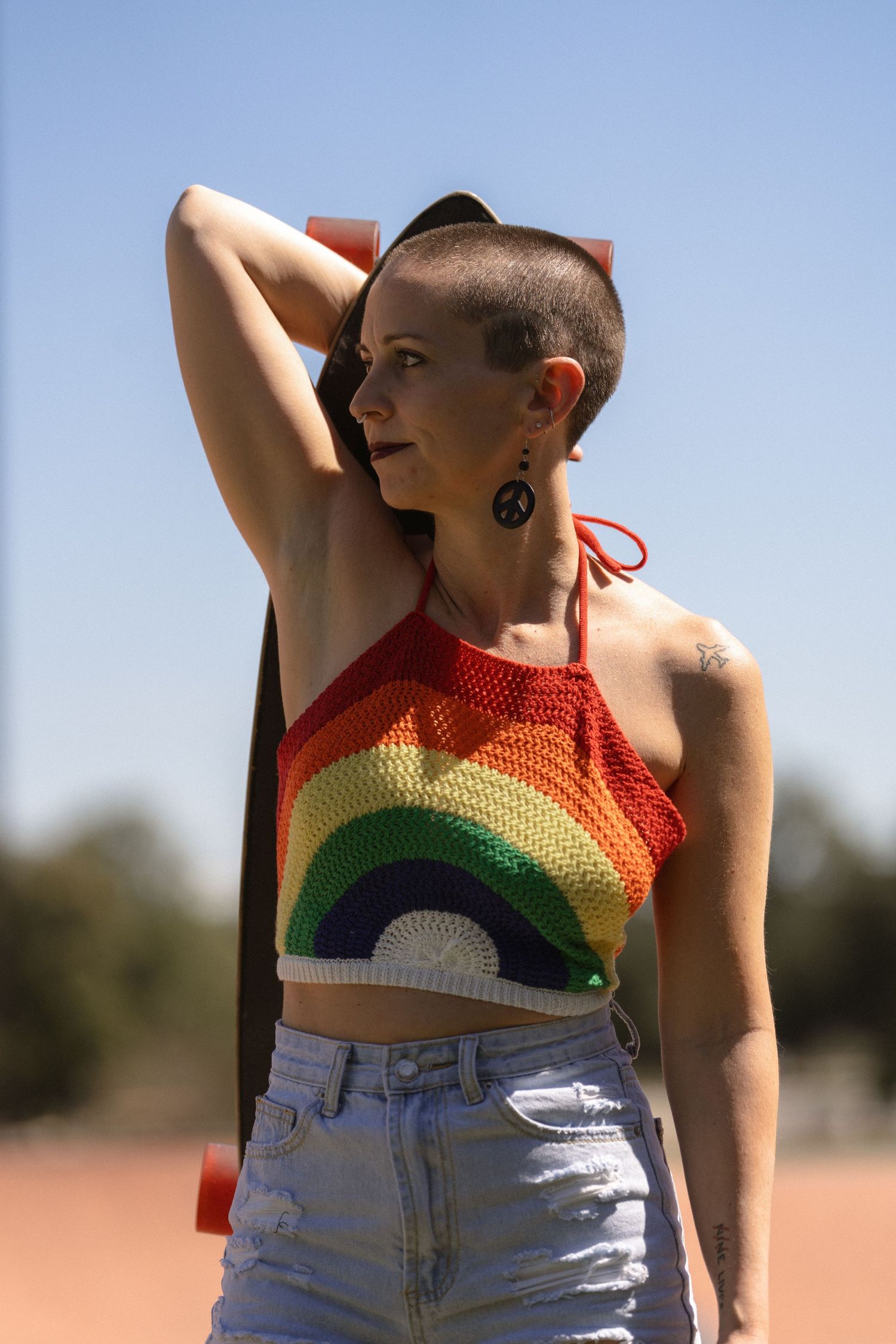A person in a crochet rainbow crop top and denim shorts poses against a bright sky.
