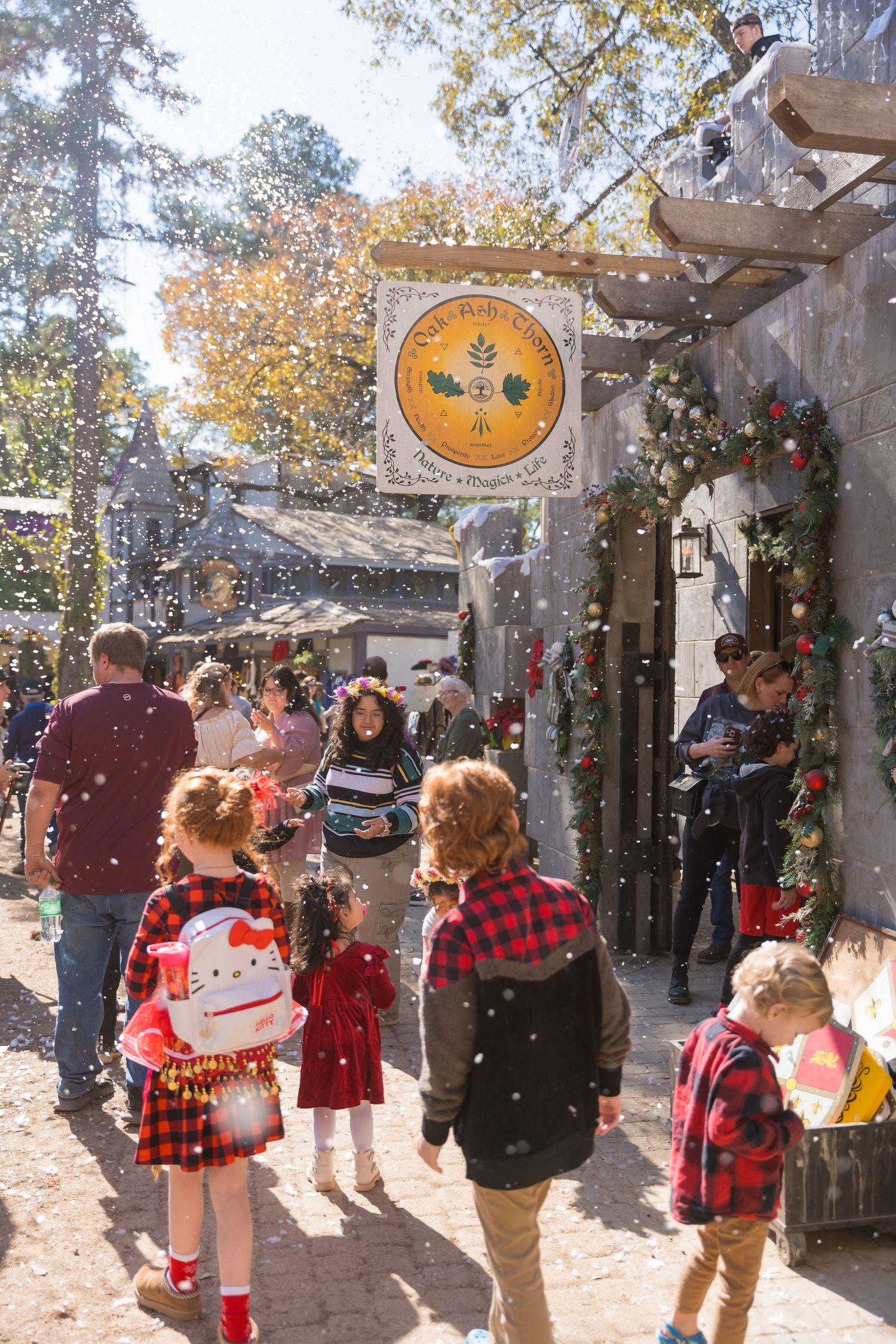 Children in festive attire walking through a decorated holiday marketplace with falling snow.