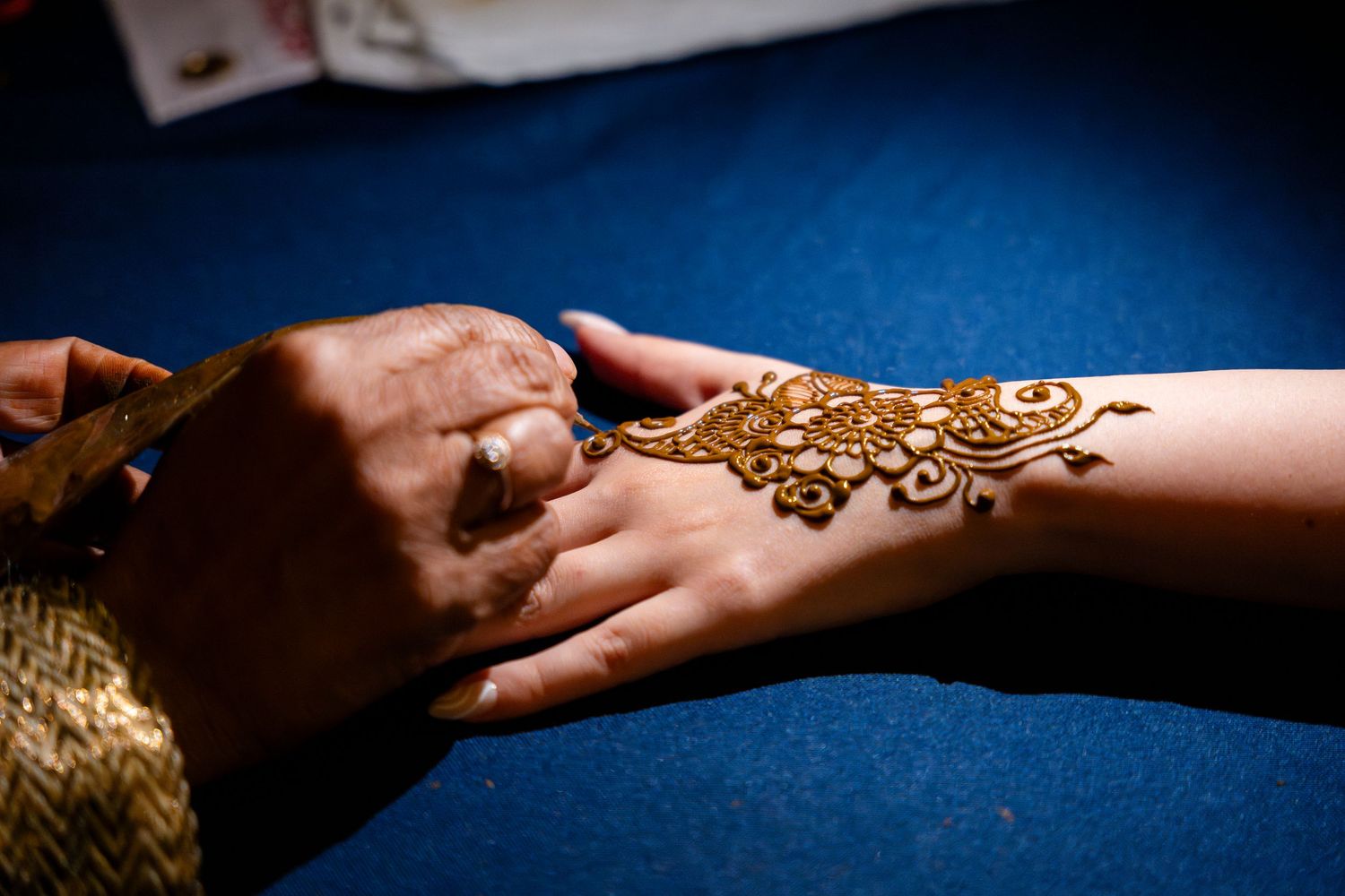 Artist carefully applies intricate henna design on person's hand against blue background in traditional mehndi ceremony.