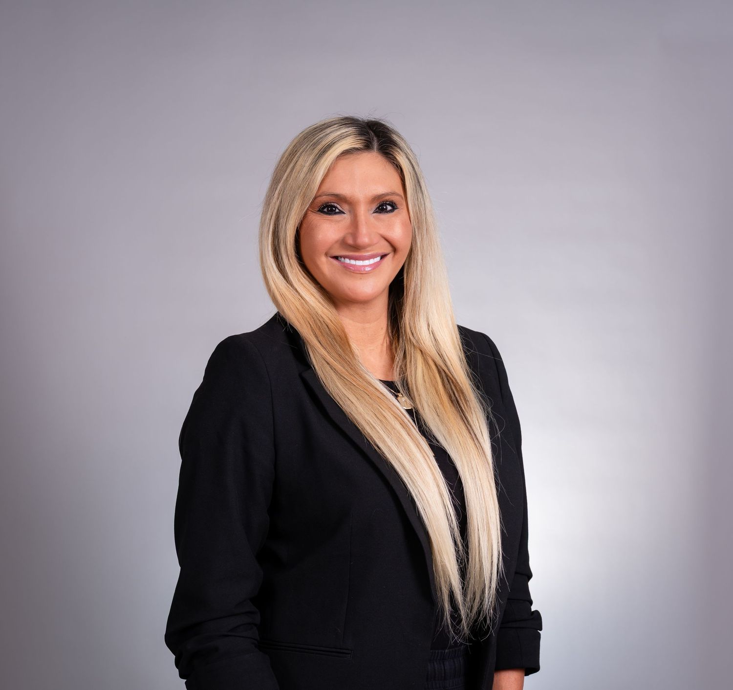 Professional corporate headshot of a person with long blonde hair wearing a black blazer against a gray background.