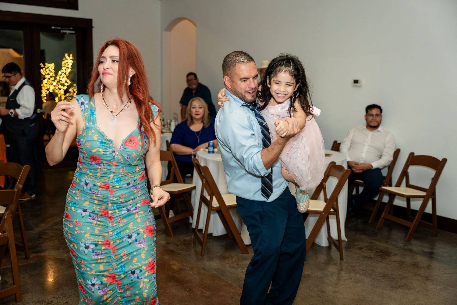 People dance together at a wedding reception in a hall with wooden chairs and polished floors.