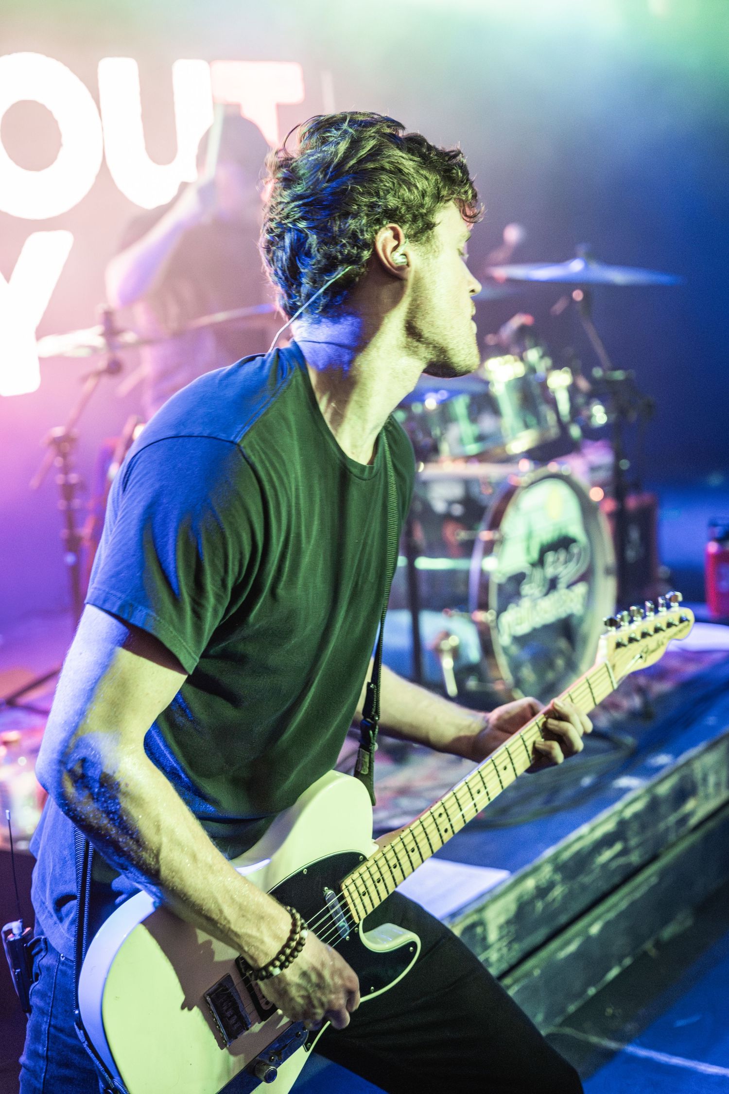 Guitarist in black t-shirt performs energetically on stage with colorful blue and pink stage lighting.
