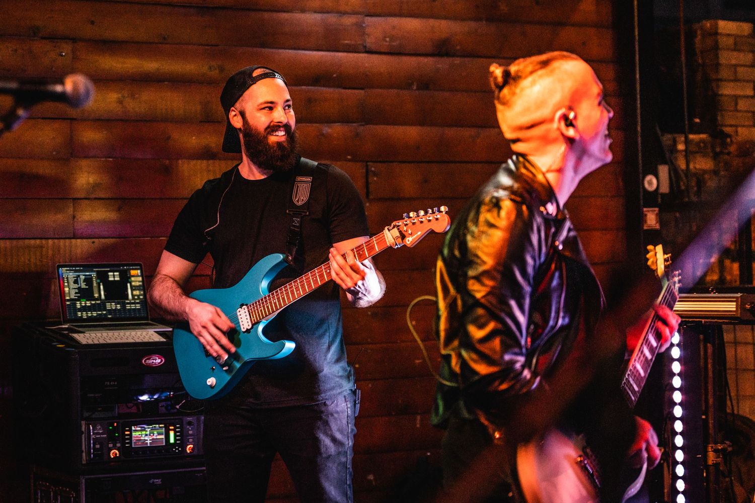 Musicians performing on stage with blue guitar against rustic wood panel backdrop.