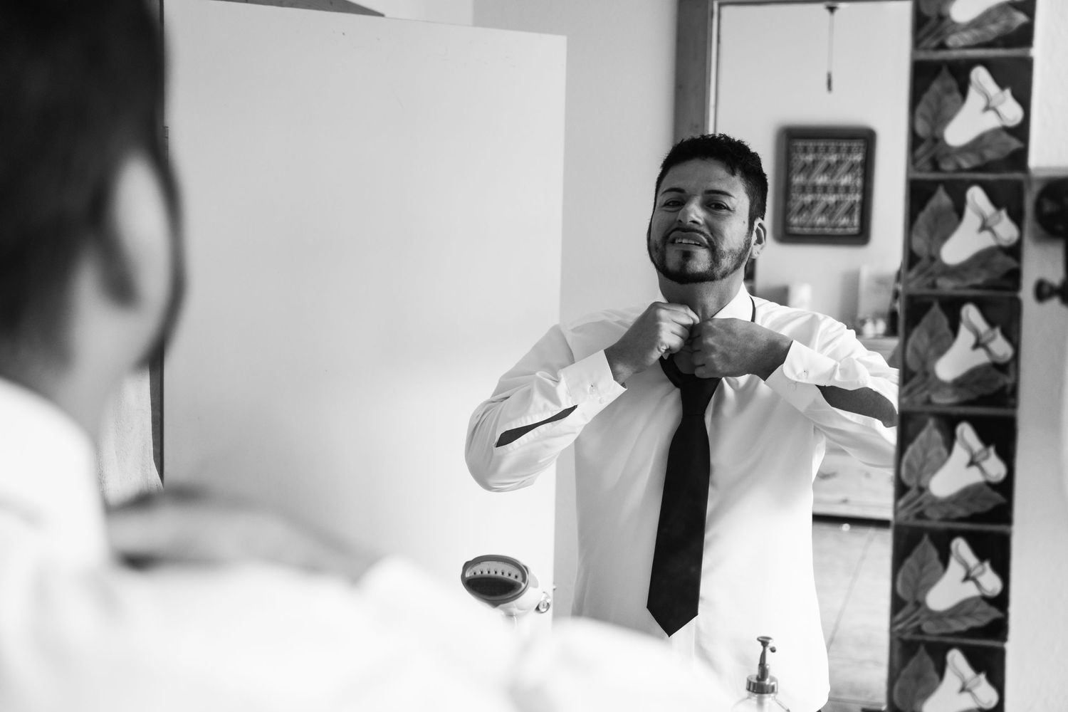 A reflection in a mirror shows someone adjusting their black tie while getting dressed in formal attire.