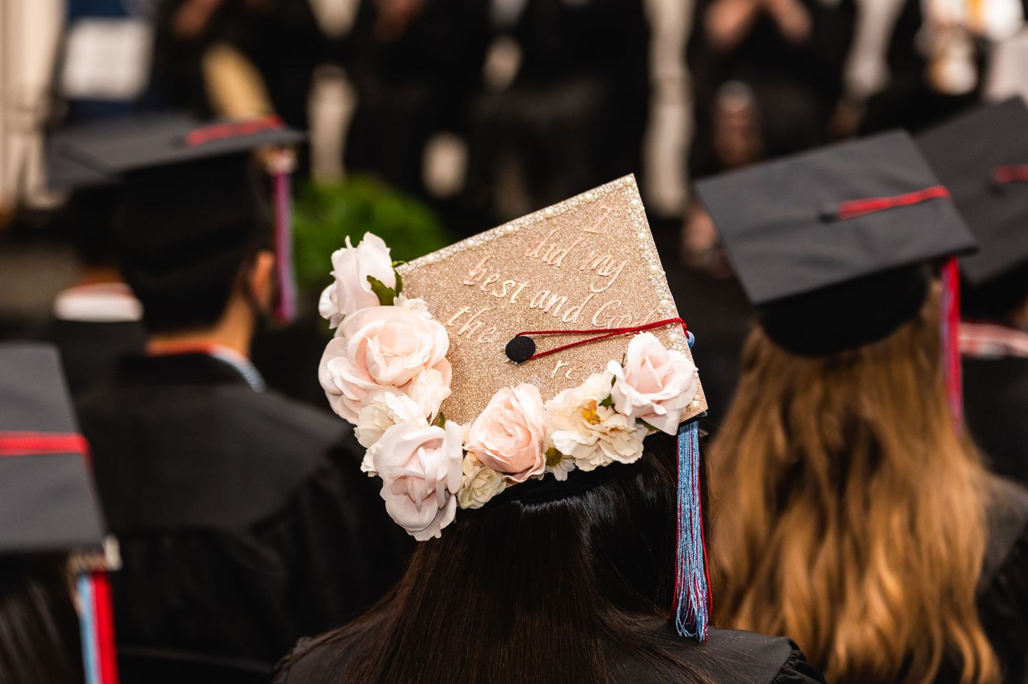 Graduation cap decorated with pink roses and tassel against blurred ceremony background.