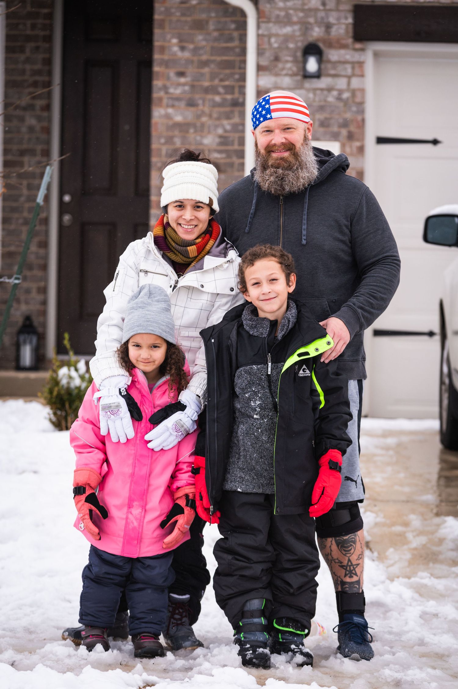 A family bundled in winter coats poses together in the snow outside their brick home.
