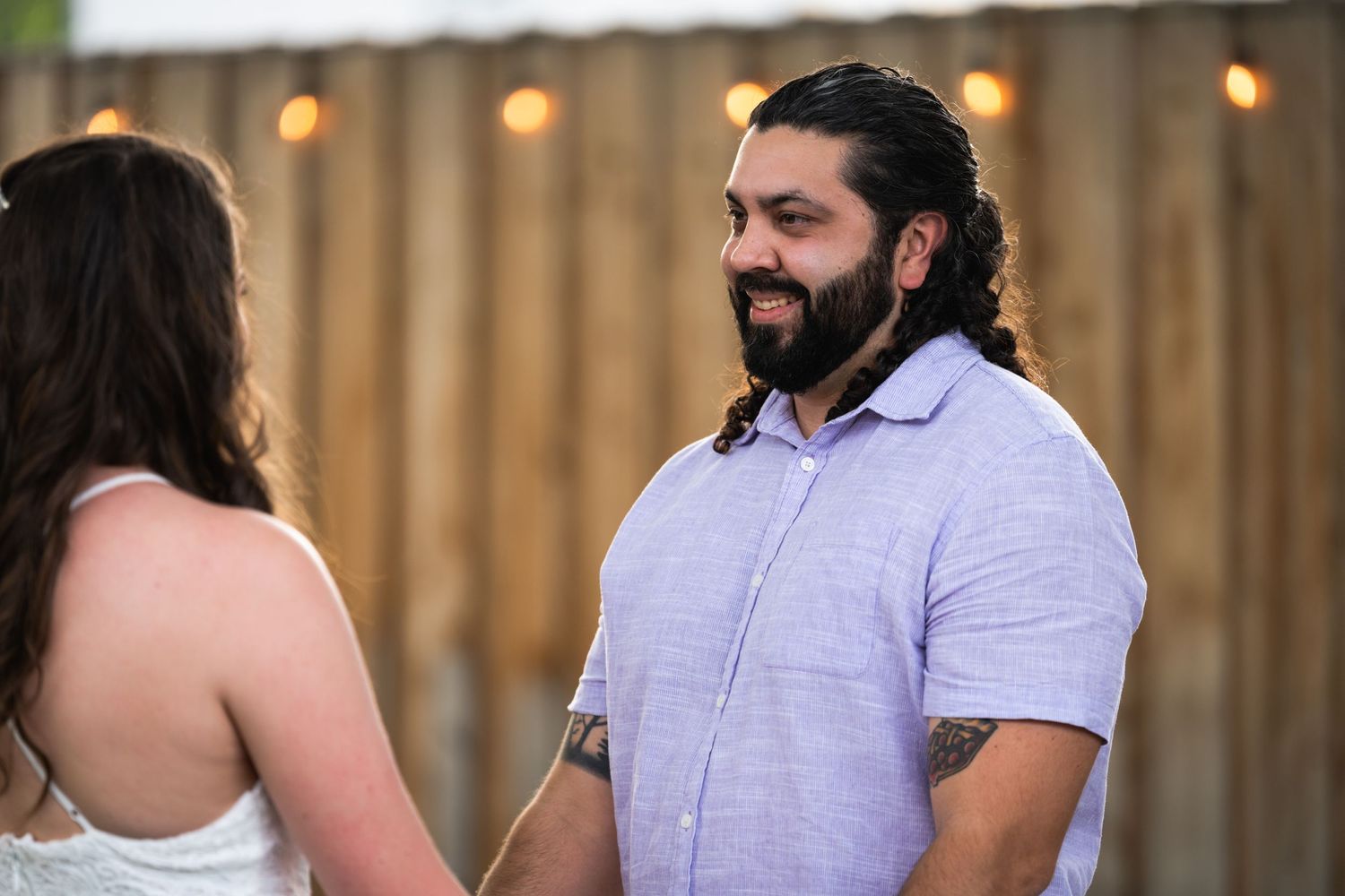 Outdoor wedding ceremony moment with wooden fence and string lights in background.