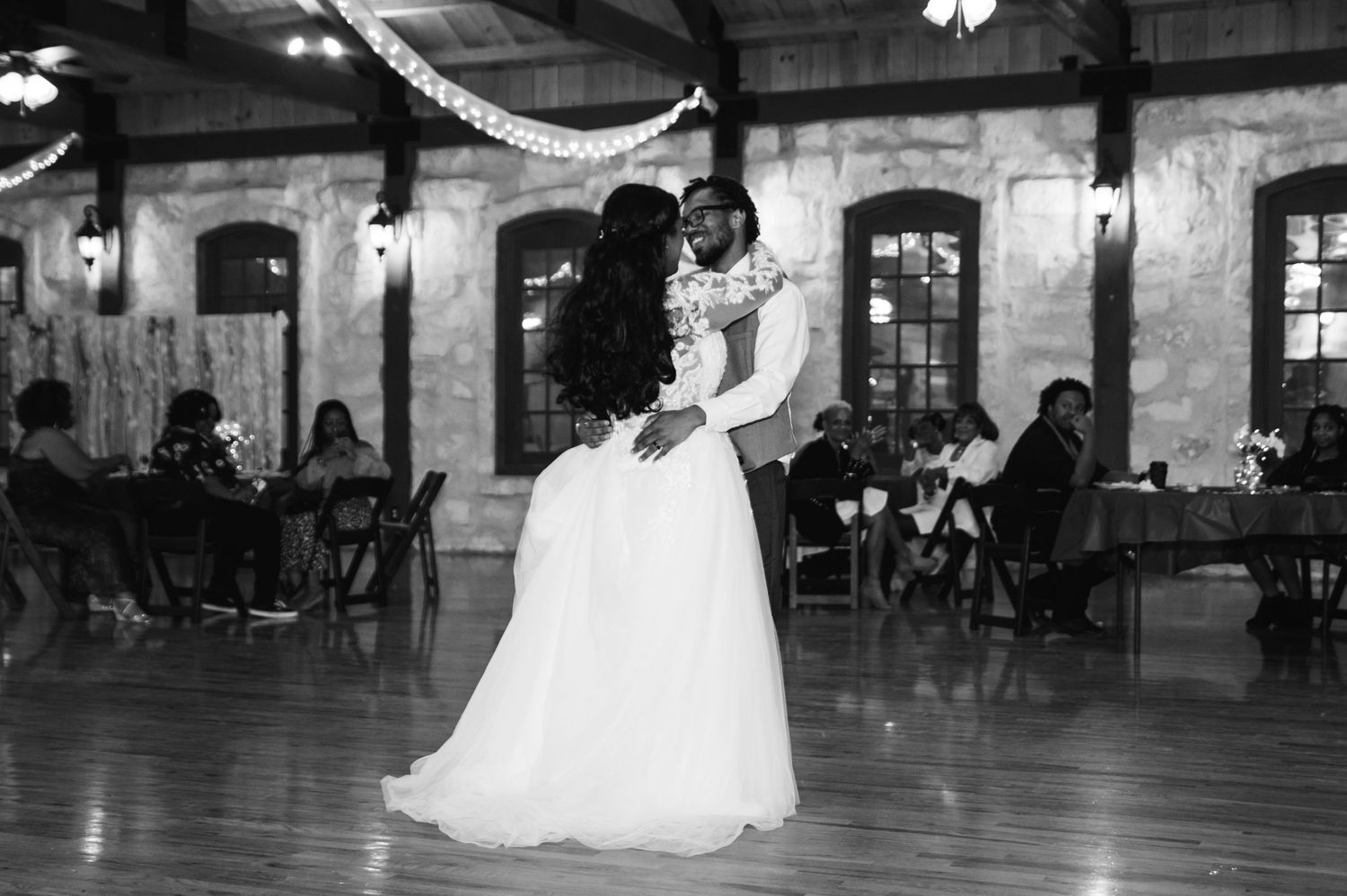 A black and white photo captures a dancing moment at an elegant wedding reception with string lights.