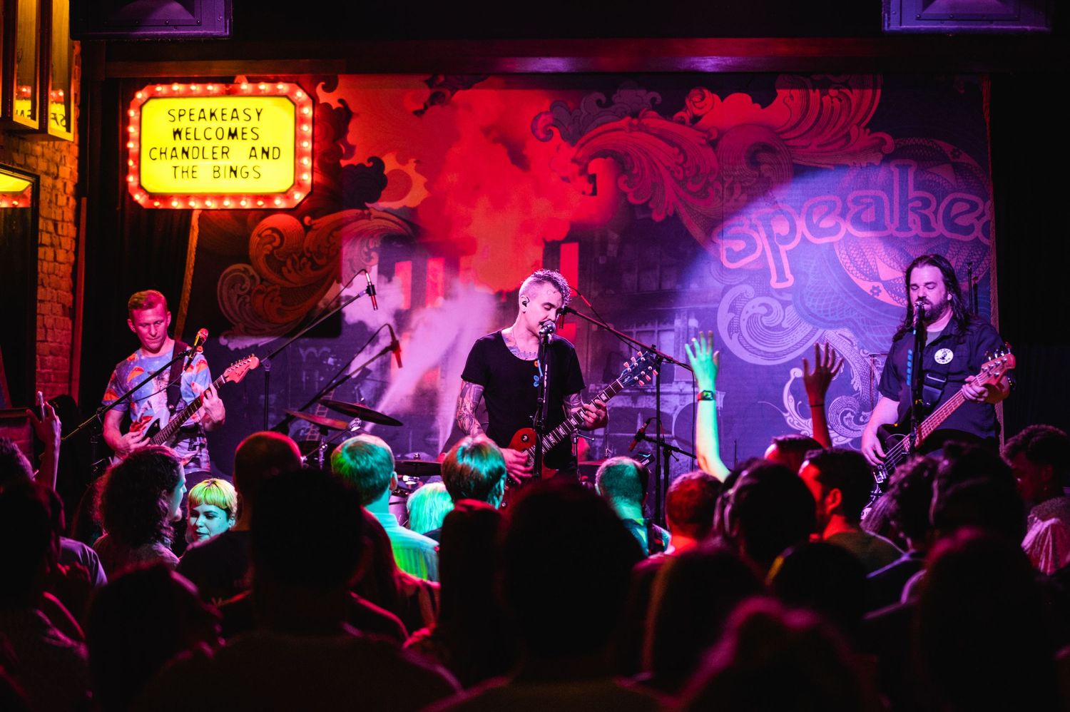 Band plays energetic show for crowd under red lighting with vintage marquee visible.
