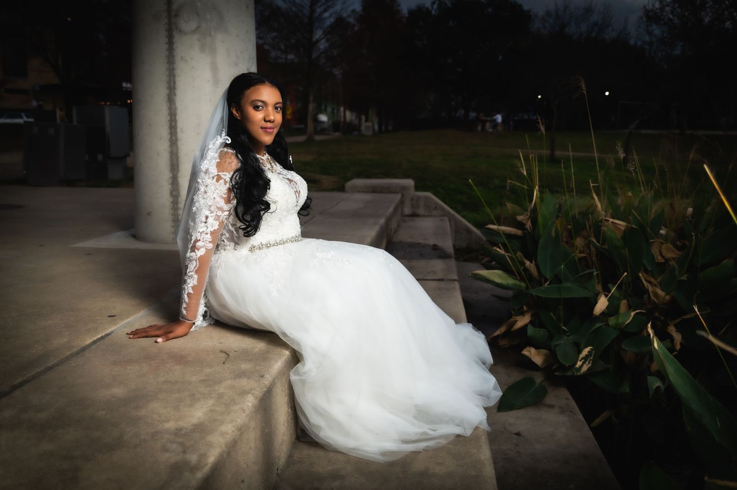 A bride in a flowing white lace dress poses on a concrete ledge at dusk with foliage in the background.