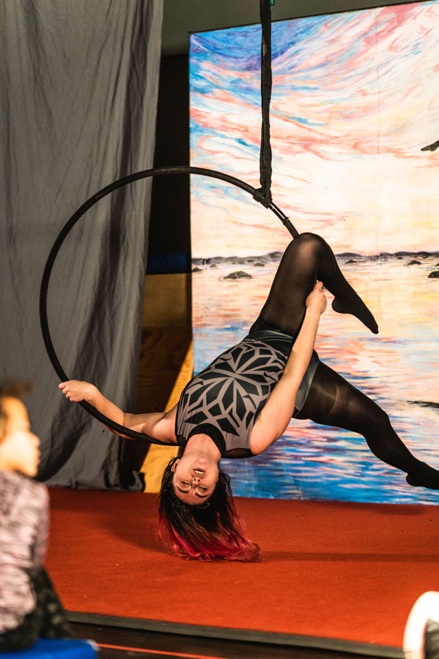 Aerial performer in black costume hangs upside down on hoop against painted sunset backdrop.