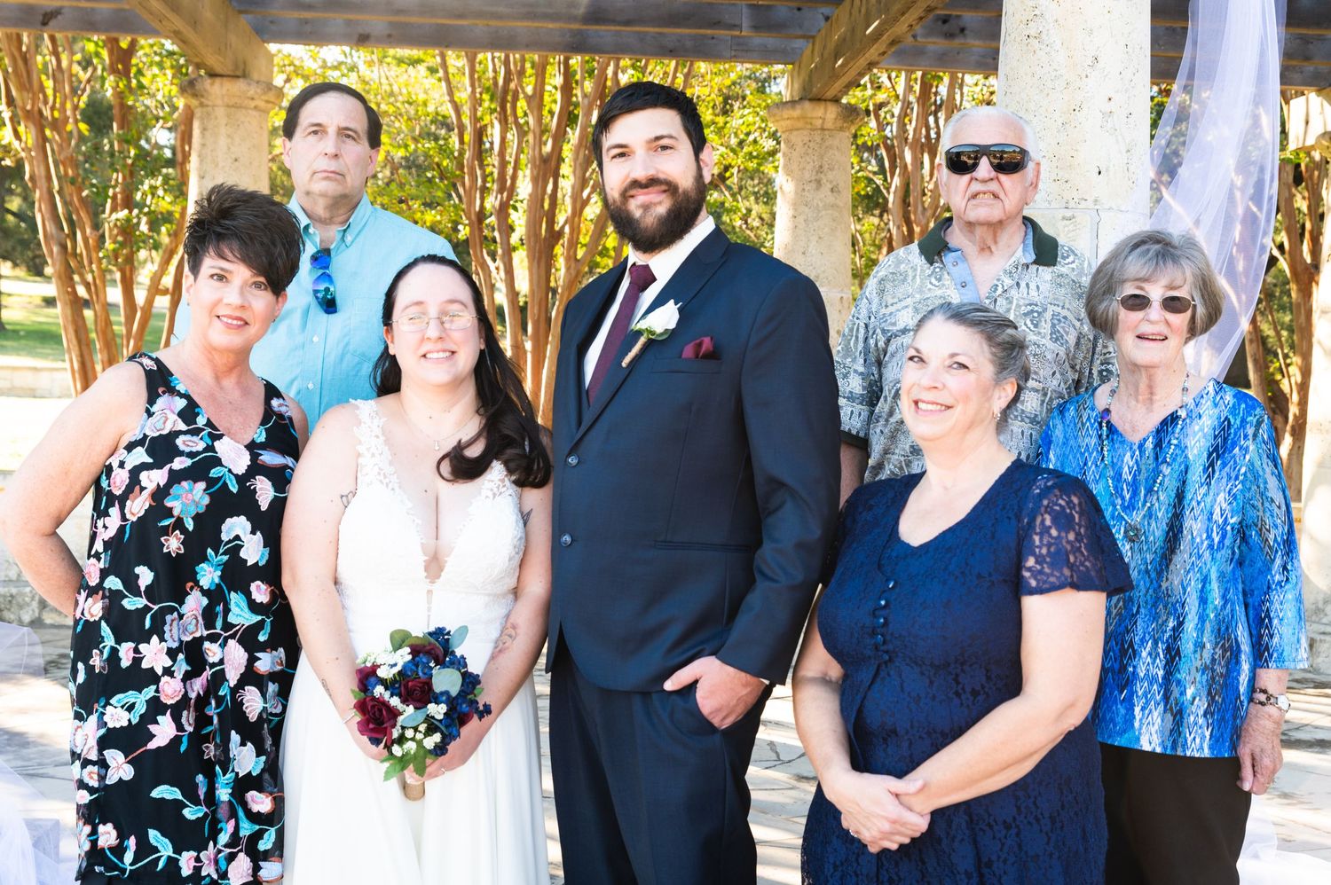 Group photo of wedding party in formal attire standing under outdoor pavilion.