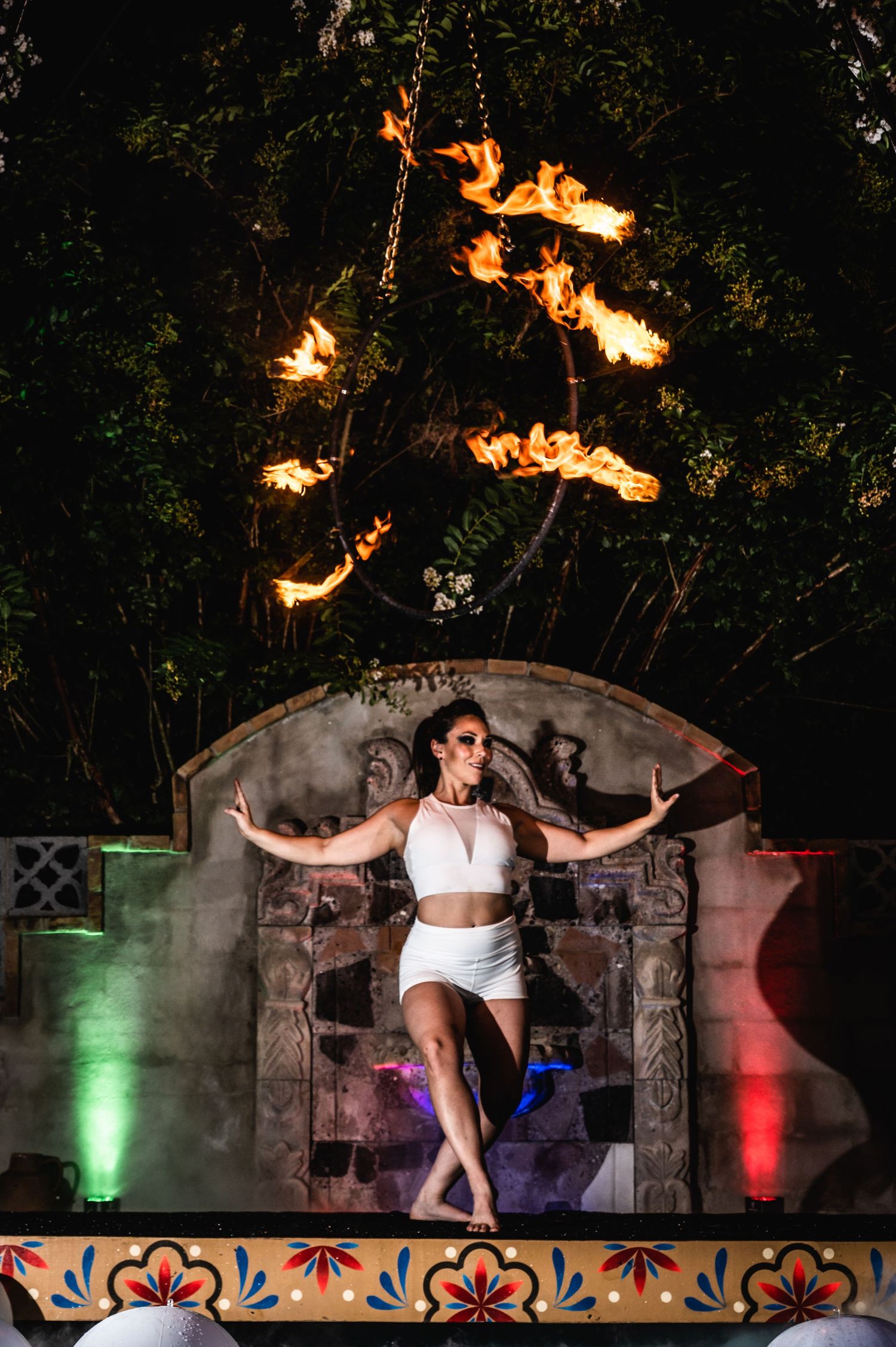 Dancer in white outfit poses dramatically against stone wall with fire elements and colored lighting.