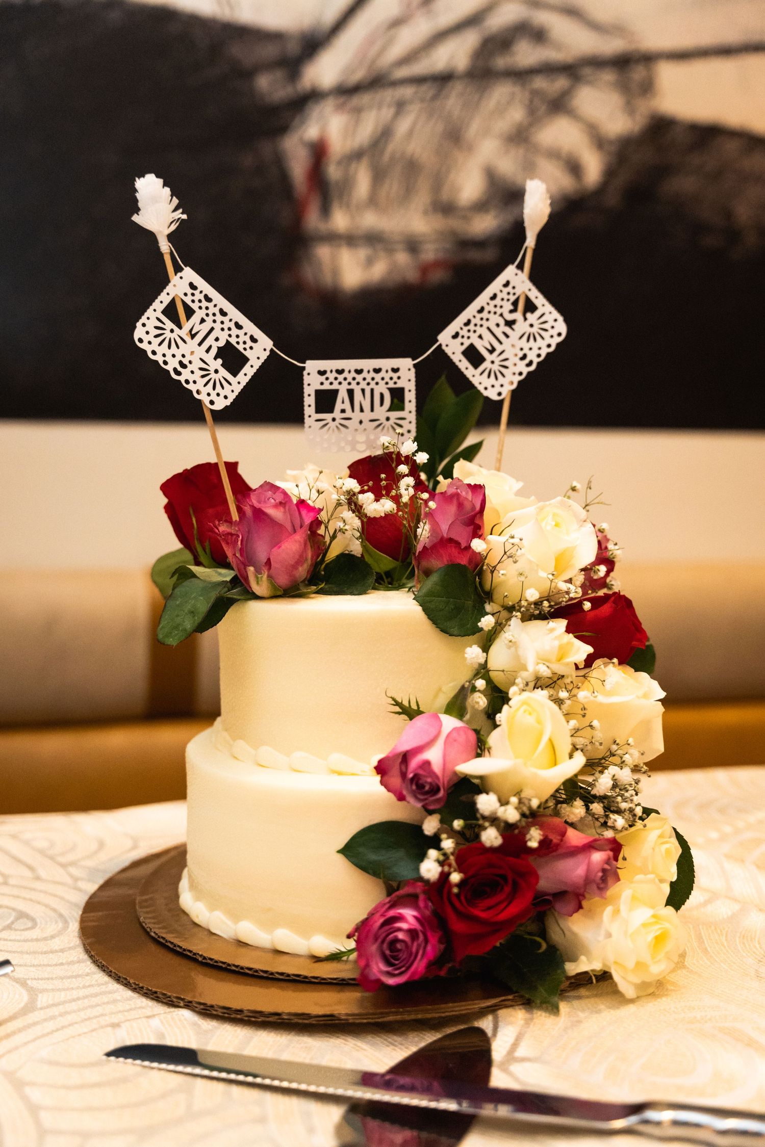 Two-tier white wedding cake decorated with pink and red roses and lace banner toppers.