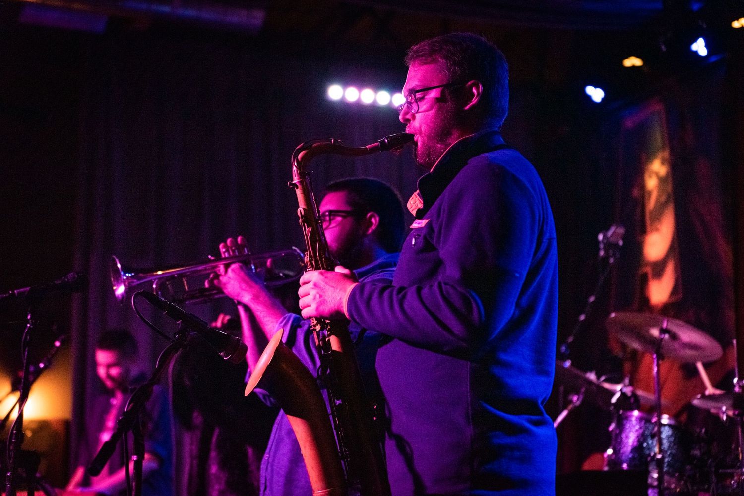 Musicians perform with brass instruments under dramatic purple stage lighting.