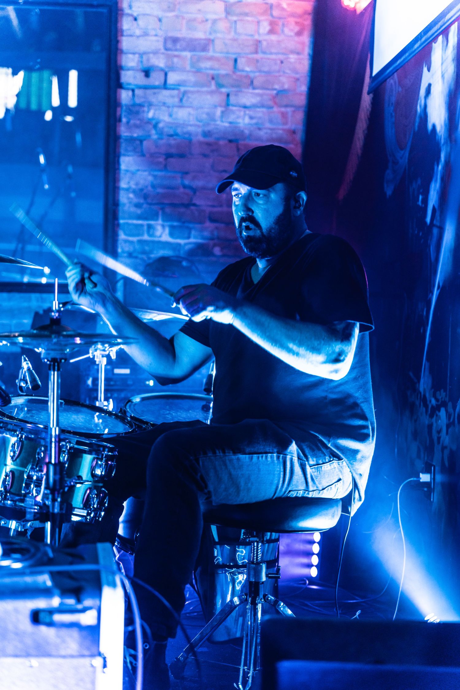 Drummer performs on stage bathed in blue lighting against brick wall backdrop.