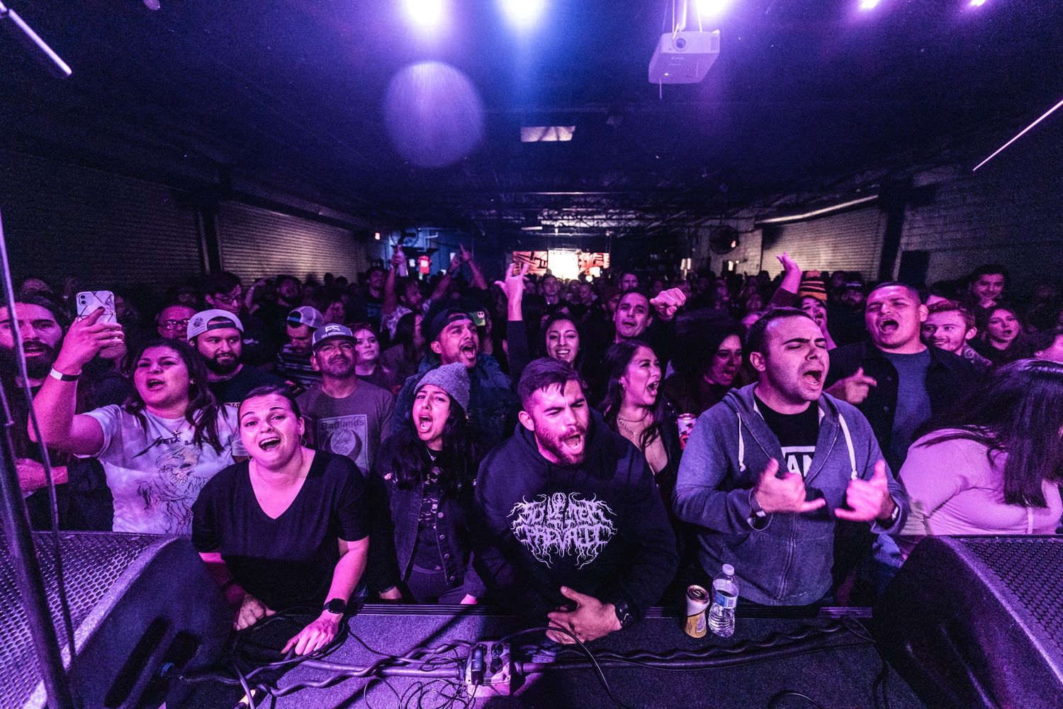 Concert crowd watches enthusiastically under purple stage lights at a live music venue.