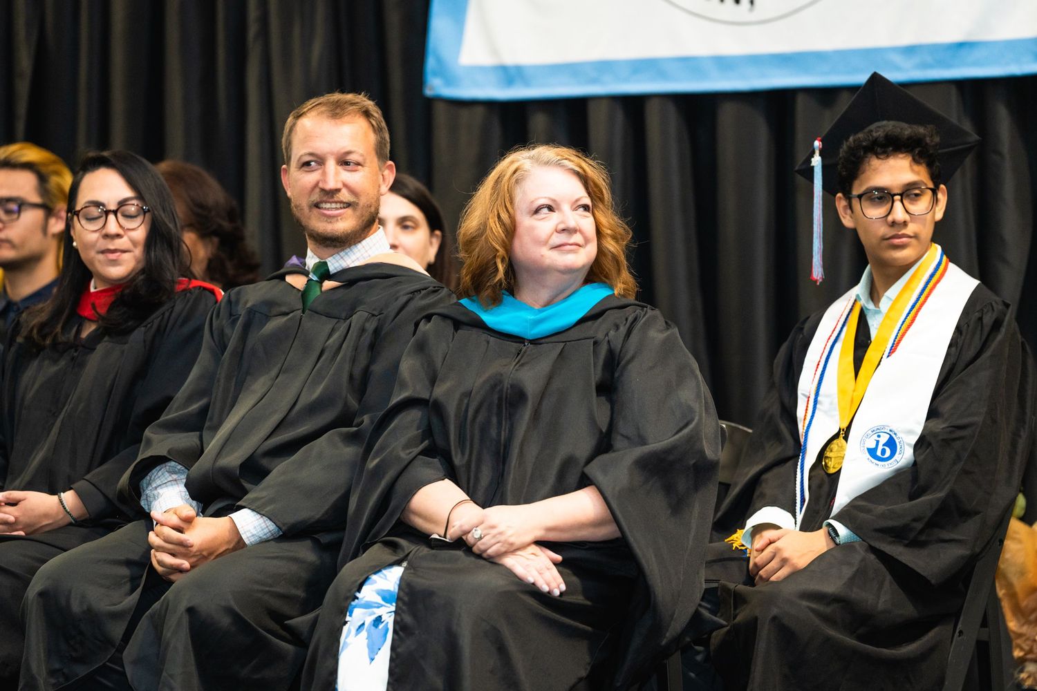 Group of graduates in traditional black academic regalia seated during commencement ceremony.
