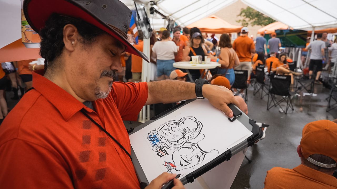 Artist in red shirt and cap sketches characters on drawing pad at outdoor event.