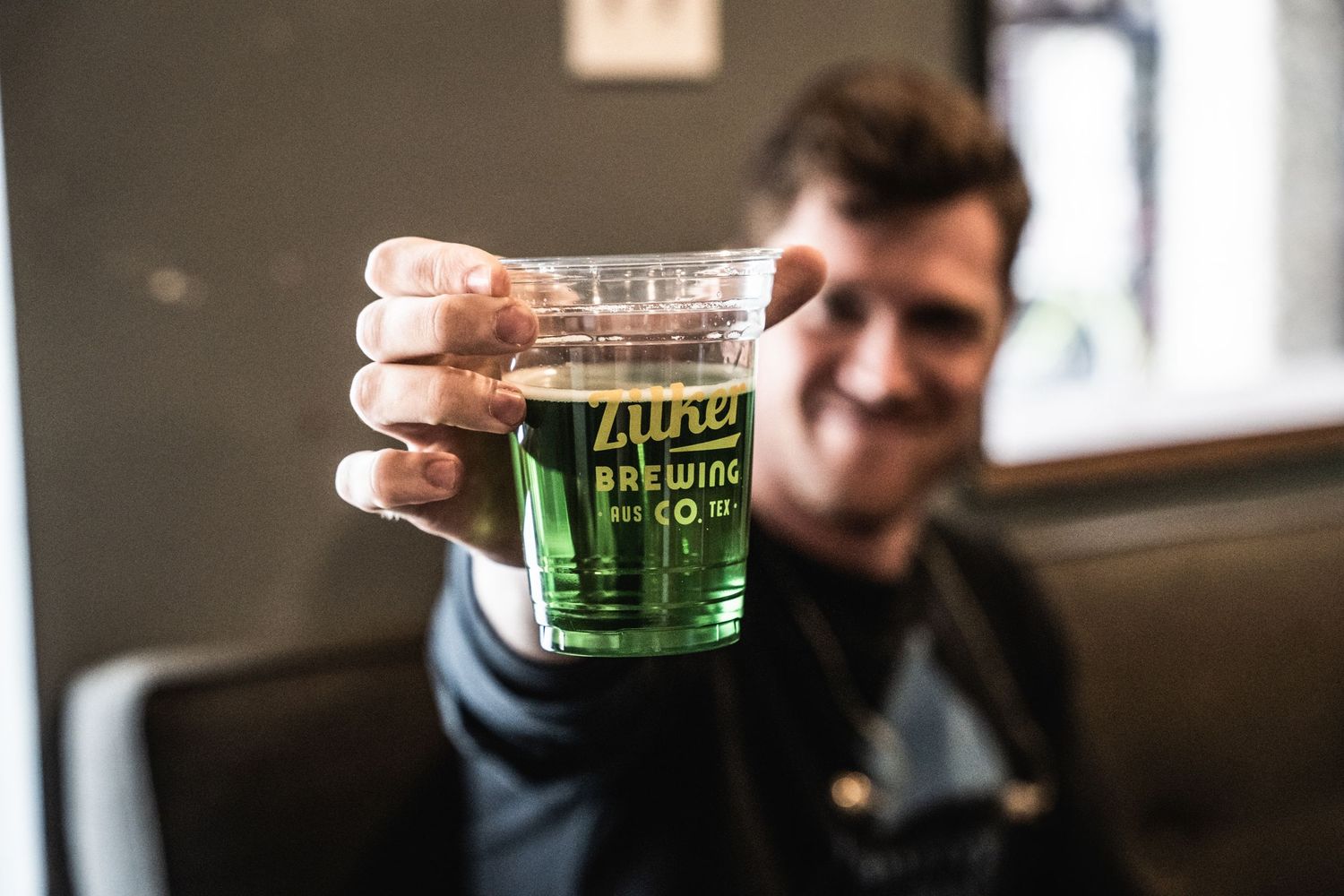 Someone holding up a glass of green beer against blurred indoor background.