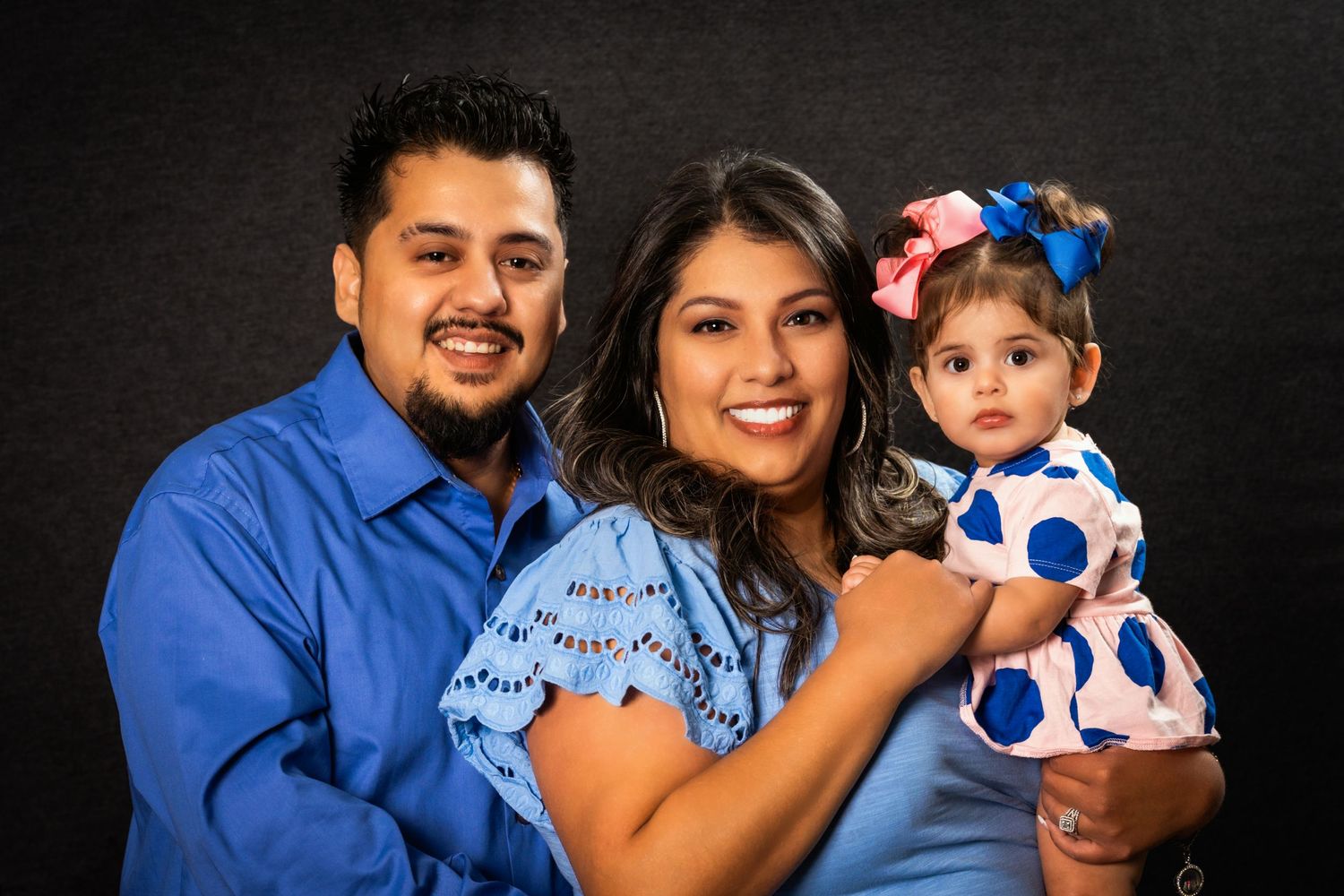 Smiling family photo with parents and baby daughter in coordinated blue and polka dot outfits.
