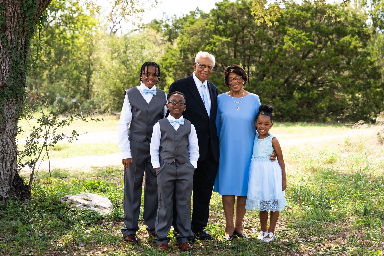 Small group in formal attire poses together under trees in natural setting.