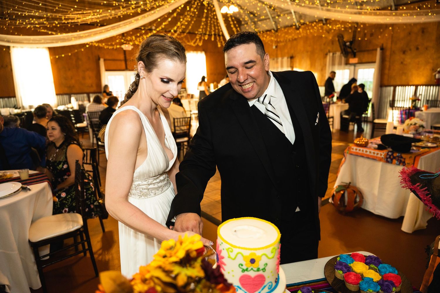 Wedding cake cutting moment under warm string lights with sunflowers and colorful decorations.
