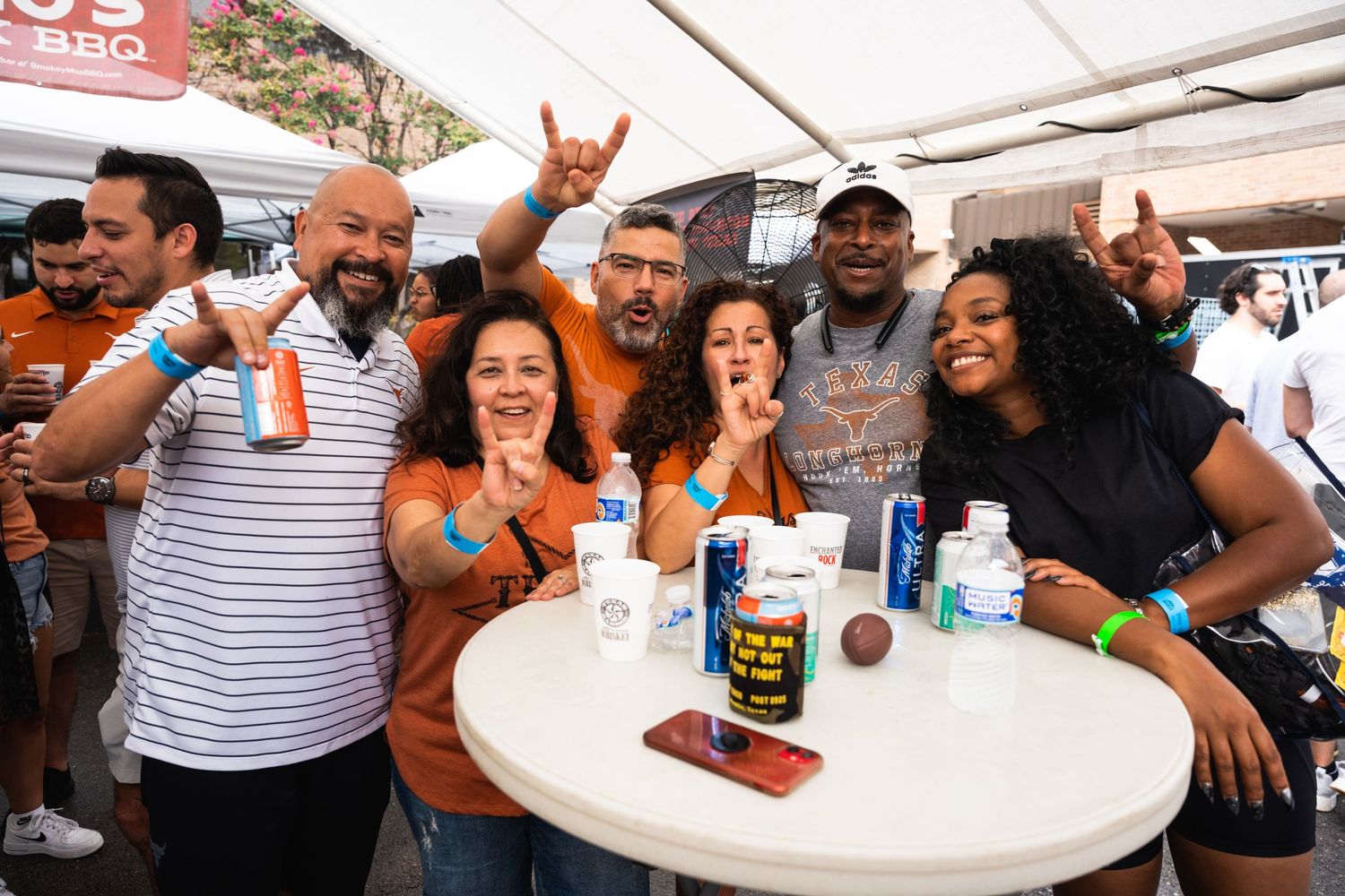 Friends making celebratory gestures around a table with drinks at an outdoor event.