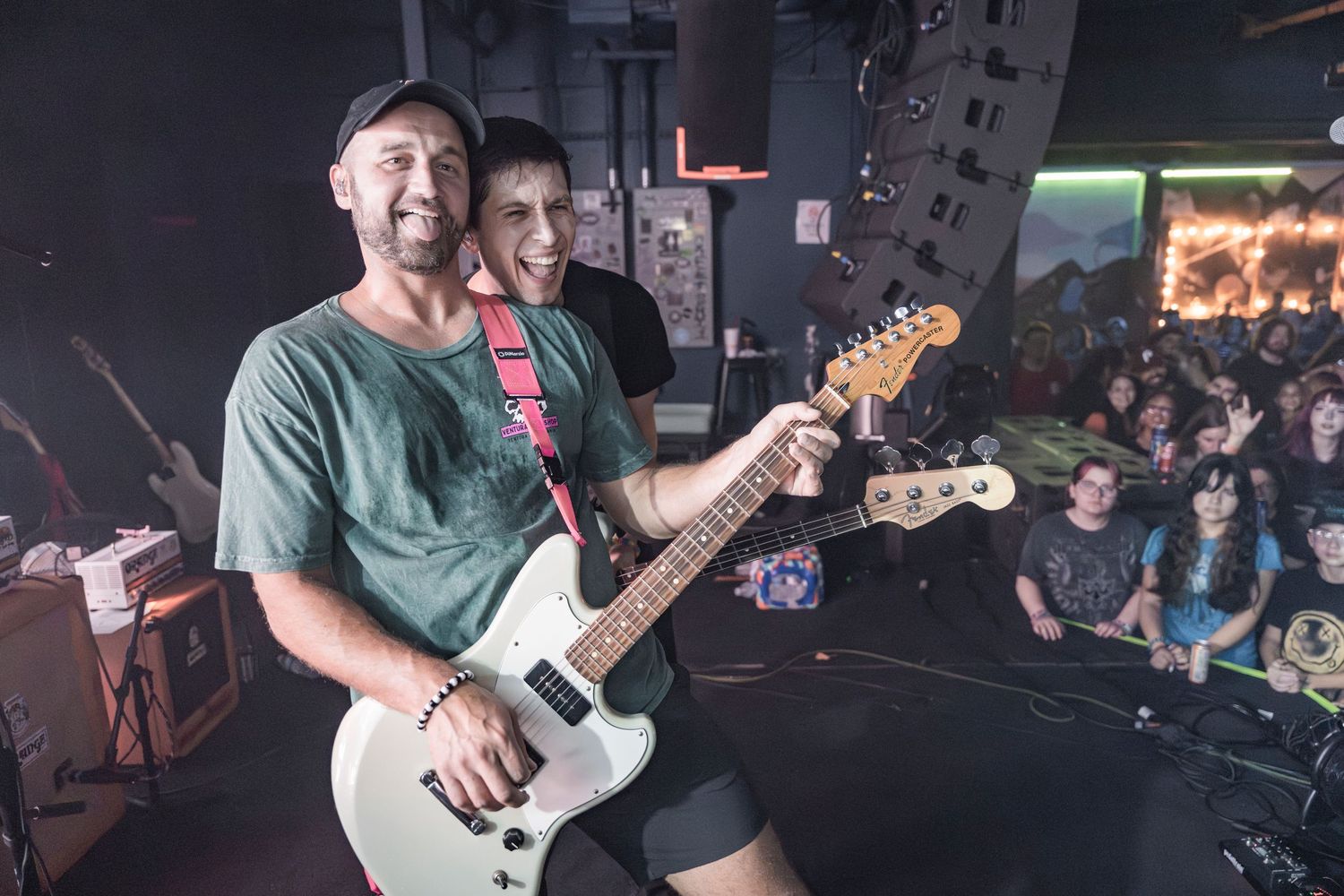 Musicians performing on stage at a live concert venue with guitars and energetic crowd in the background.