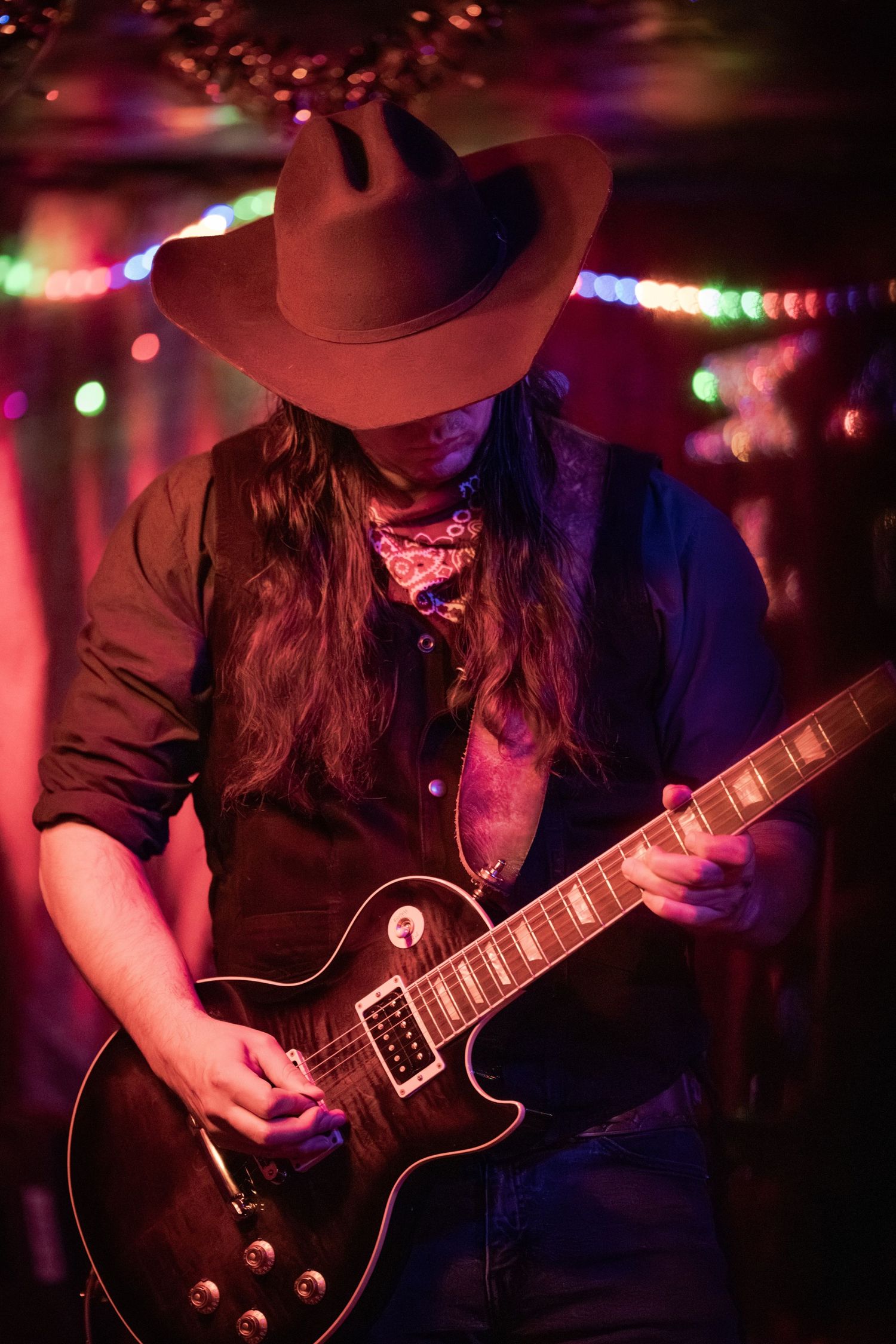 Electric guitarist performs on stage with a black guitar under colorful stage lighting in a dark venue.